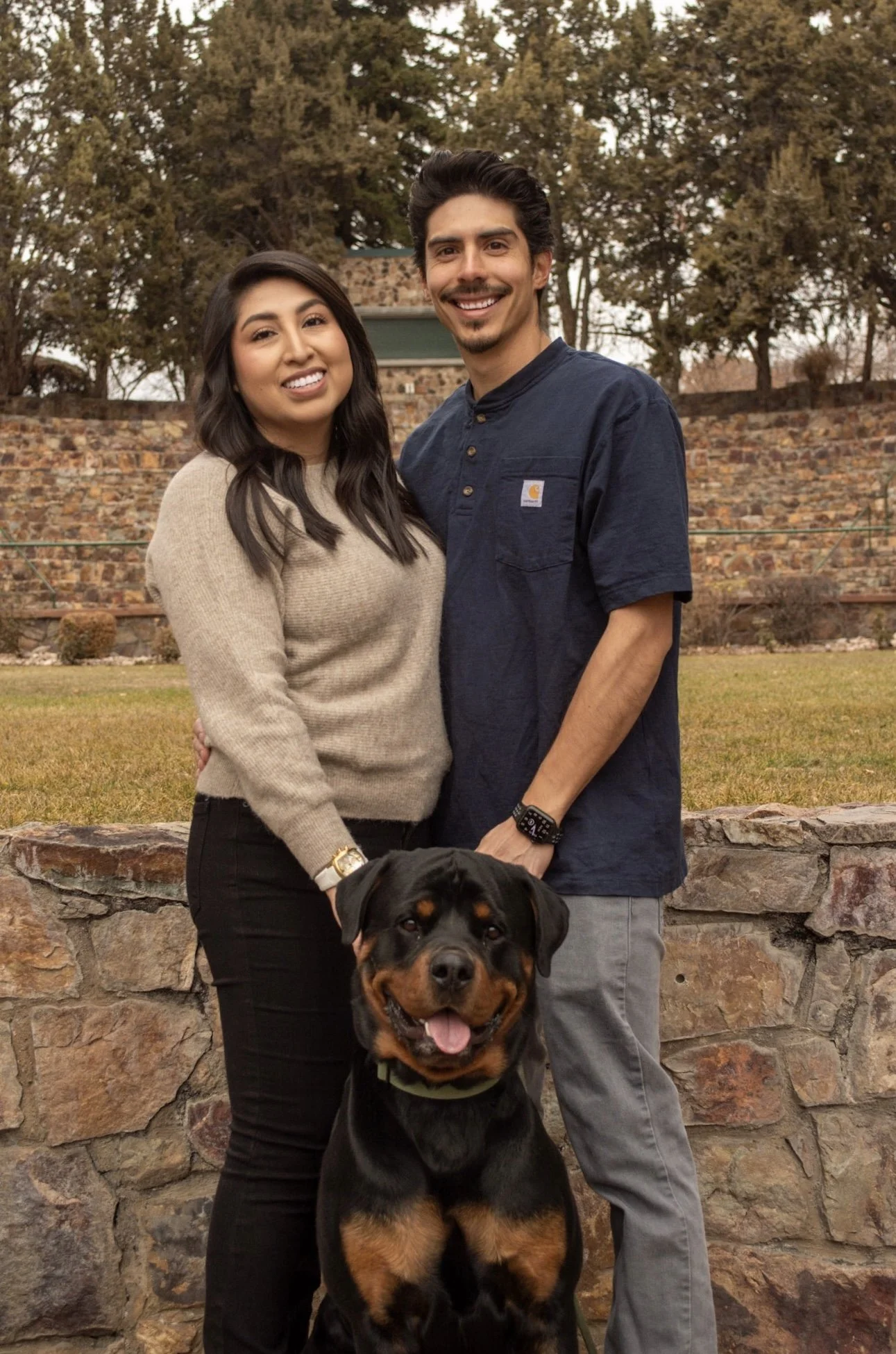 A smiling couple standing outside in front of a stone wall and trees, with a Rottweiler dog sitting in front of them.