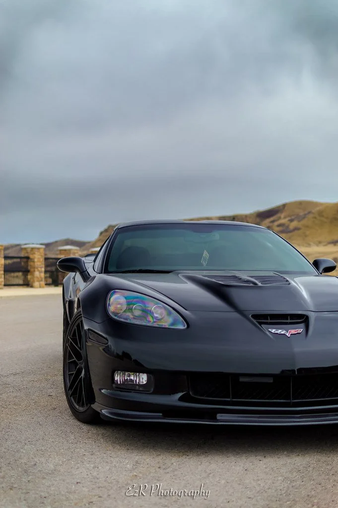 A black Chevrolet Corvette sports car parked outdoors with a cloudy sky and hilly landscape in the background.