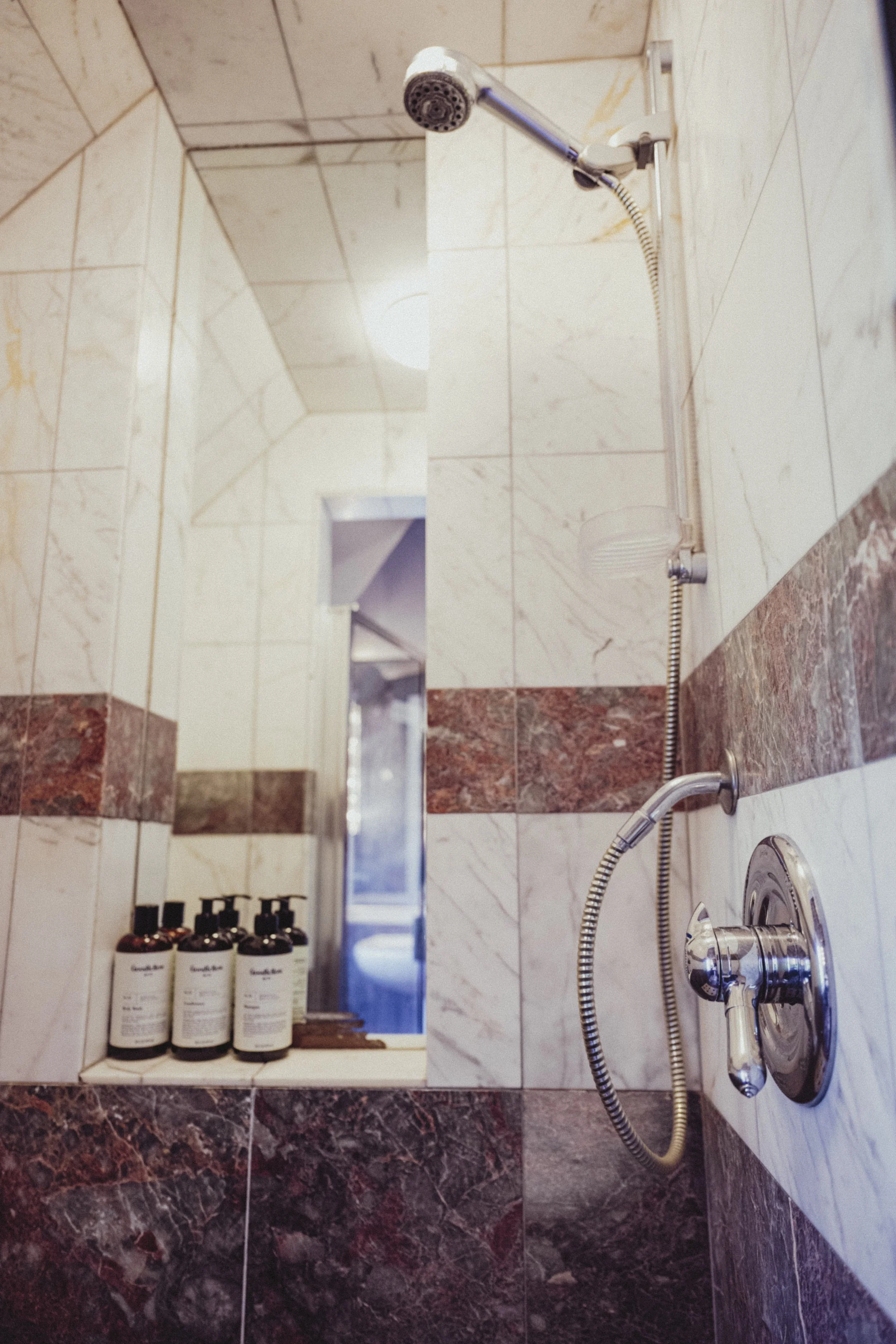 View of a shower area with a mirror, bottles of shampoo or body wash, and shower fixtures, including a showerhead and a handheld shower wand, on tiled walls.