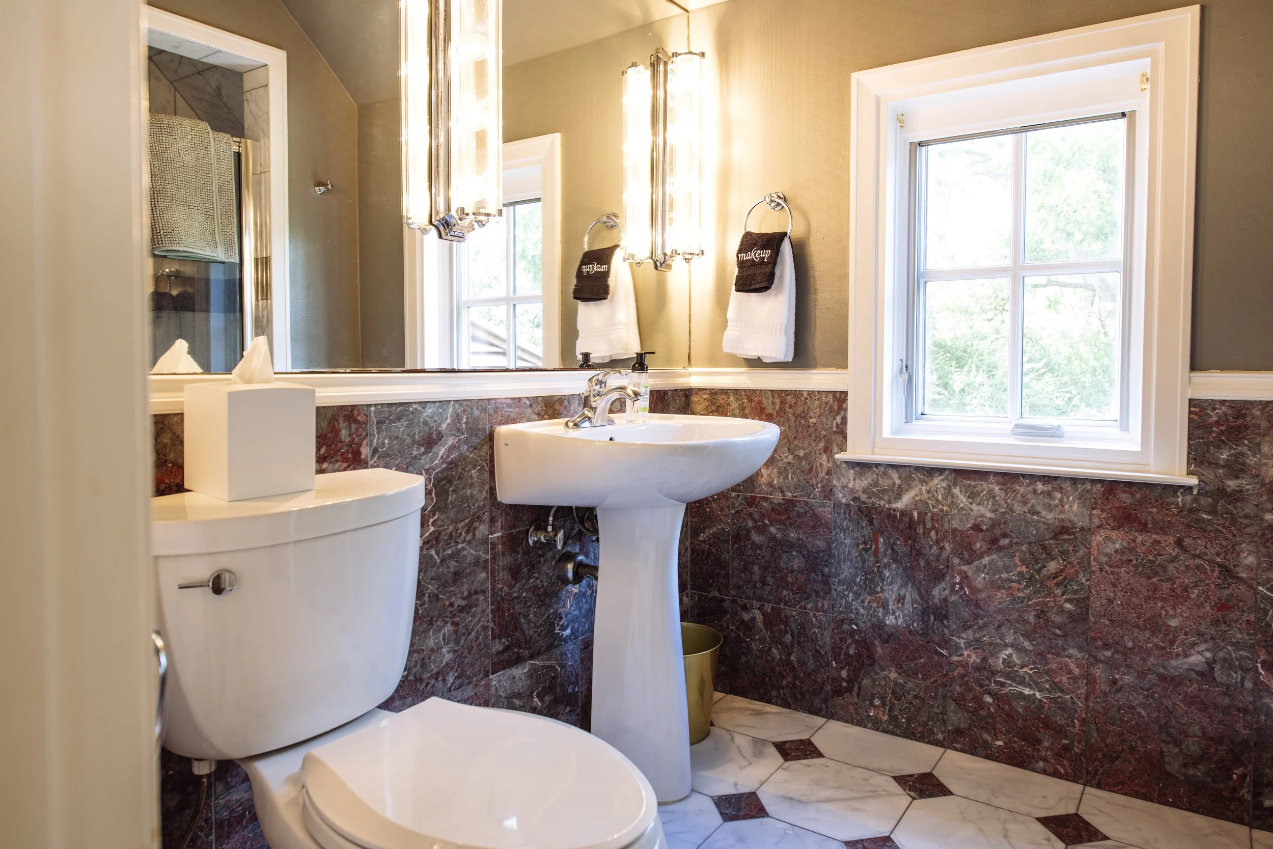 A small, elegant bathroom with a toilet, a white pedestal sink, a large mirror, wall-mounted lights, and a window with a view of trees outside. The walls are painted beige, and the lower half is decorated with red and gray marble tiles. Hand towels h