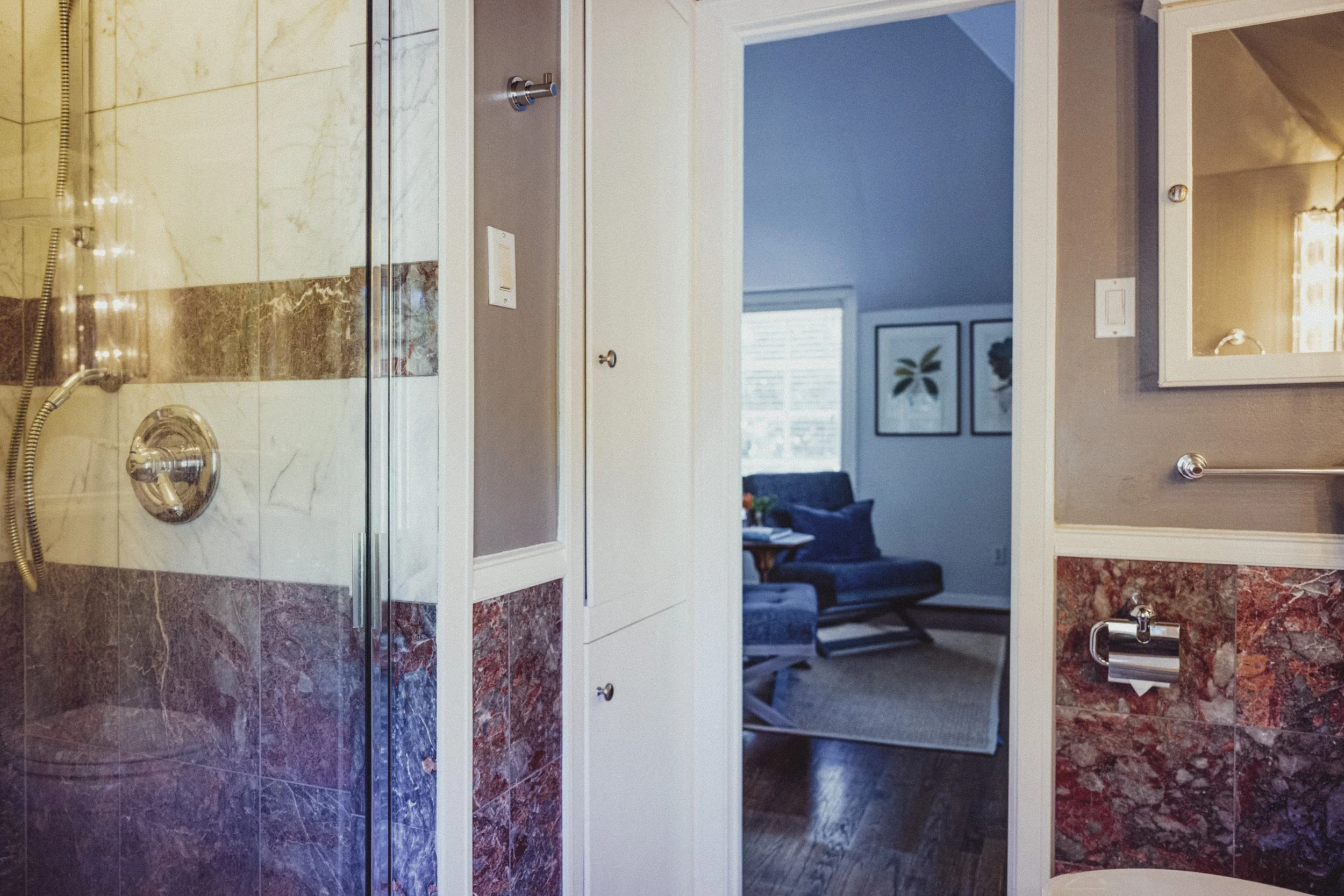 View from a bathroom showing a glass shower door on the left, with tiled walls in beige and maroon marble. The bathroom wall is painted gray with a mirror and chrome towel rack on the right. Through the bathroom doorway, a living room with large wind
