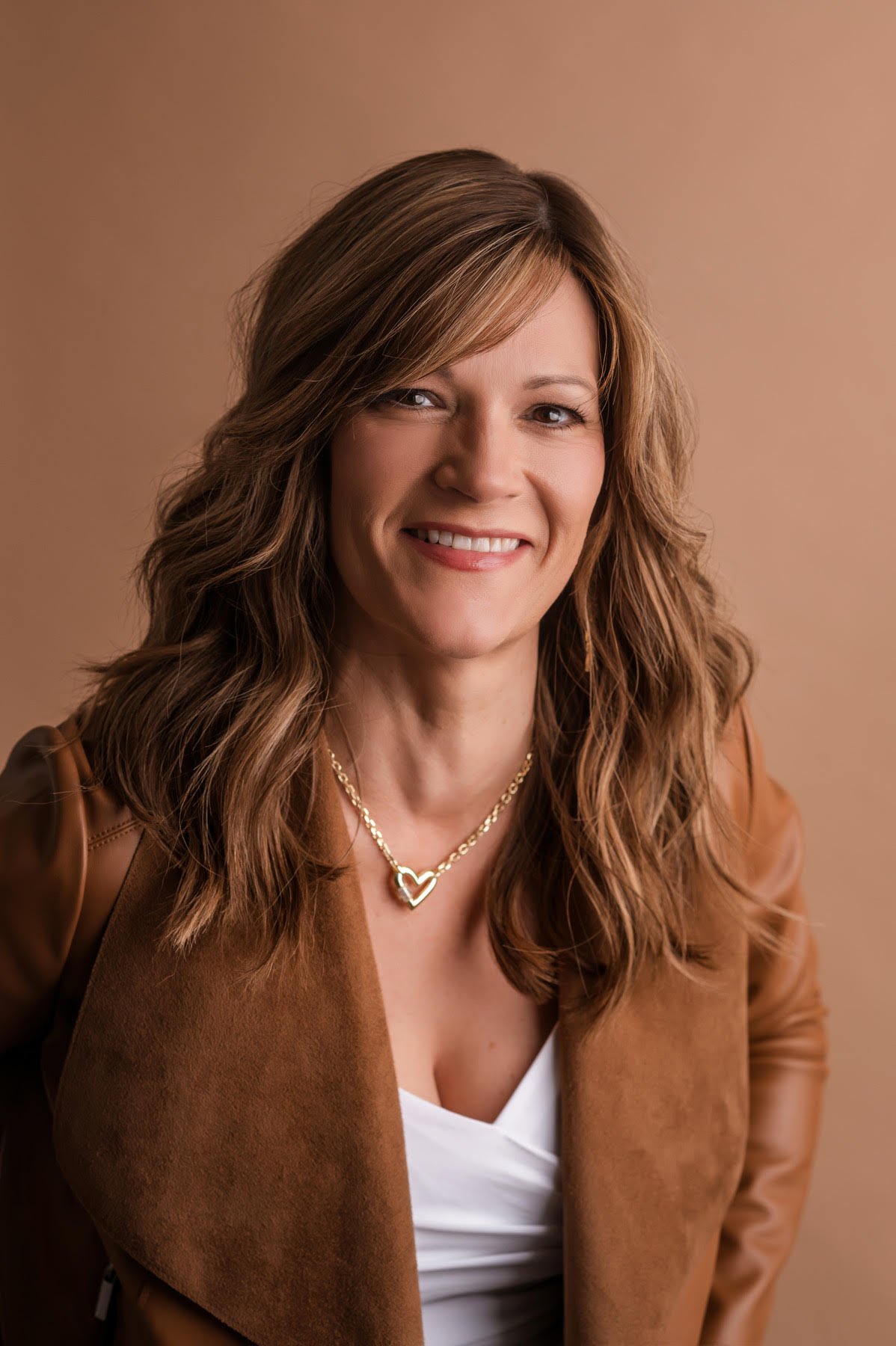 Independent stylist at an Elkhorn, Nebraska salon sitting in a modern white studio wearing a soft pink dress and smiling. Specializes in blonding, color correction, and advanced hair loss solutions.
