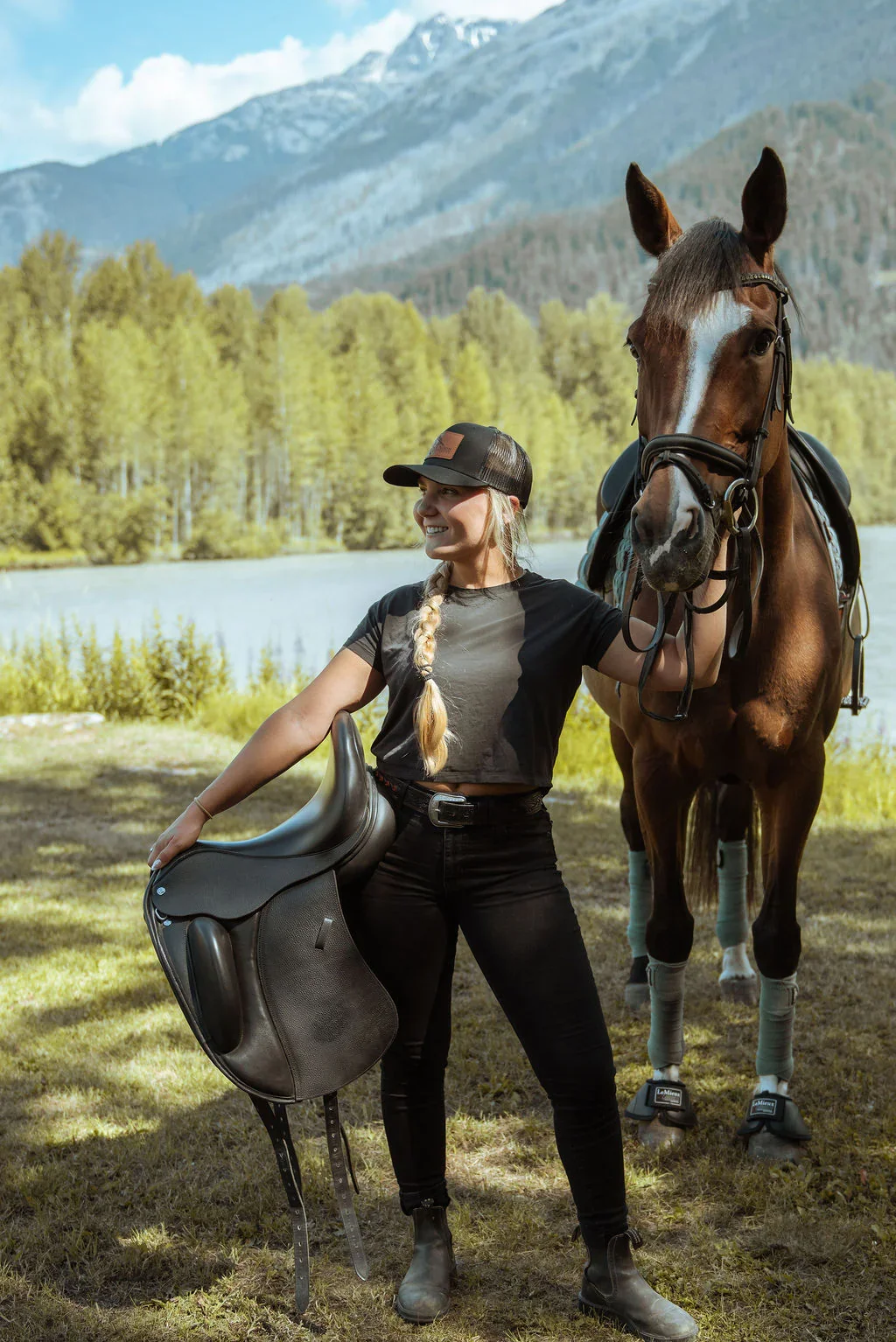A woman in black riding attire stands outdoors next to a brown horse with leg wraps, holding the horse's bridle. She is smiling, wearing a black cap, and holding a black saddle pad. The background features a lake, green trees, and mountains under a partly cloudy sky.