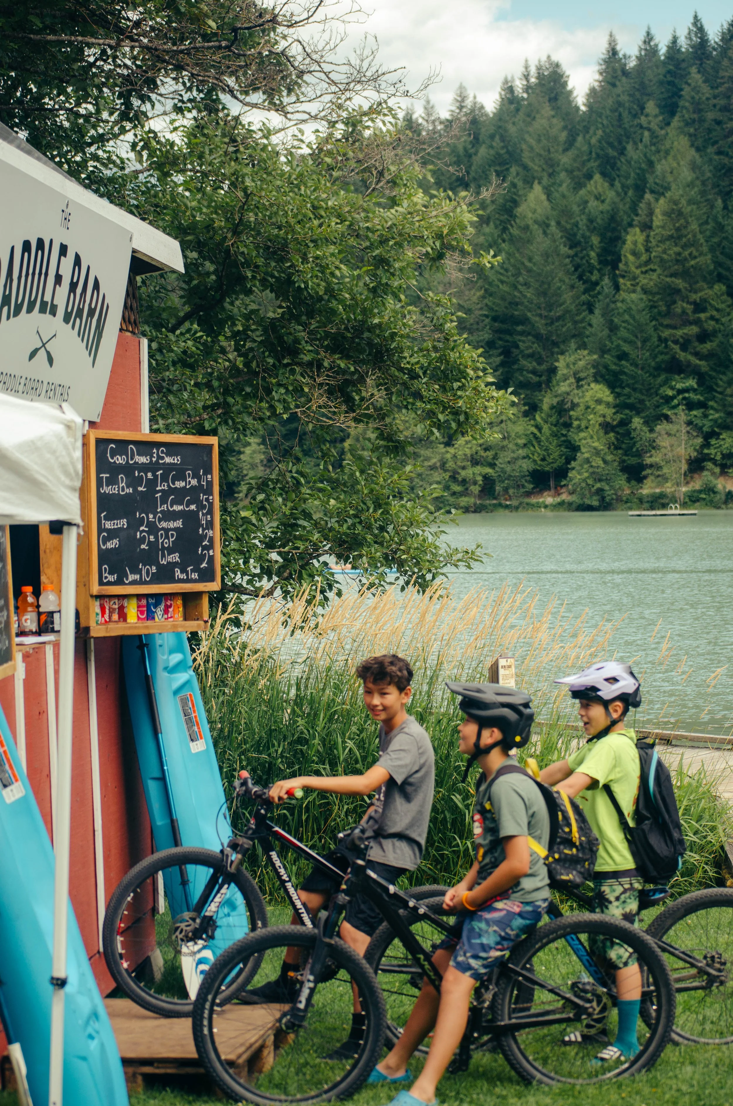 Three boys with bicycles near a lakeside stand with paddleboards and a menu board, surrounded by trees and water.