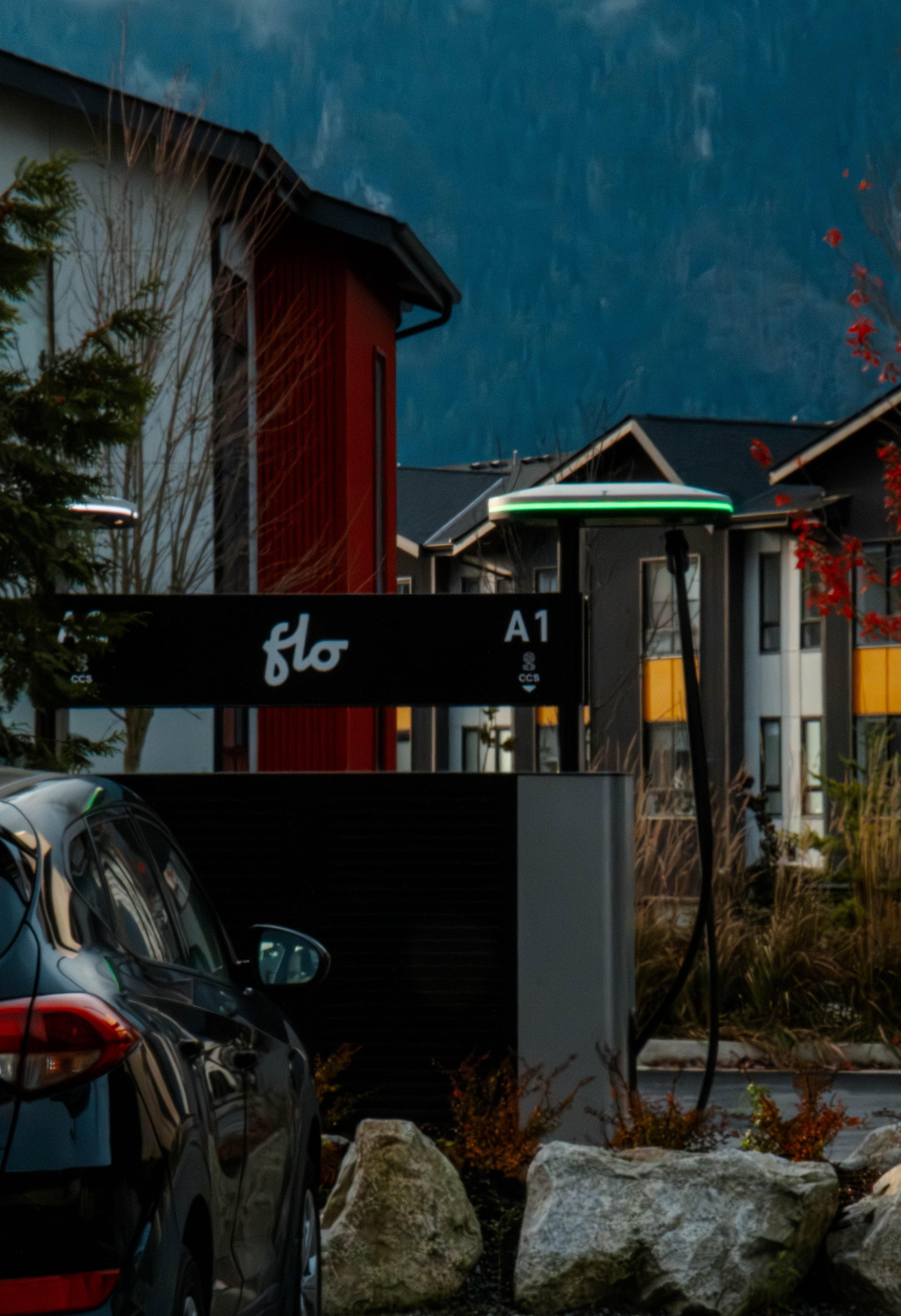 An electric vehicle charging station next to a black car in a residential neighborhood with modern houses and trees, with mountains in the background.