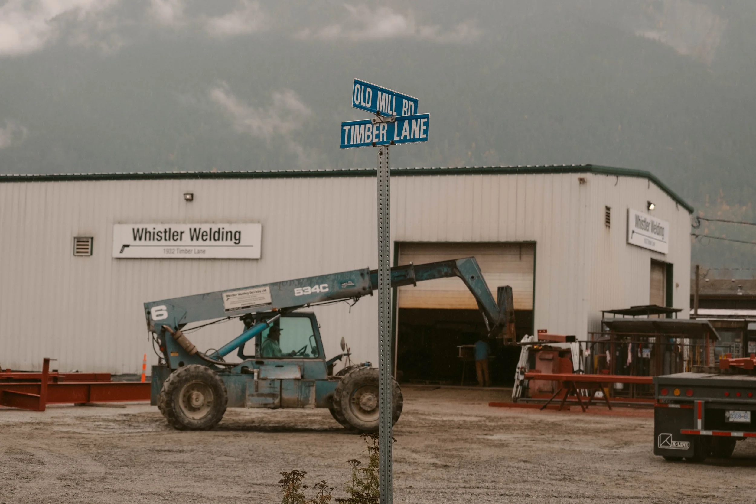 Street sign at the corner of Old Mill Road and Timber Lane, with a warehouse building, construction equipment, and trucks in the background.