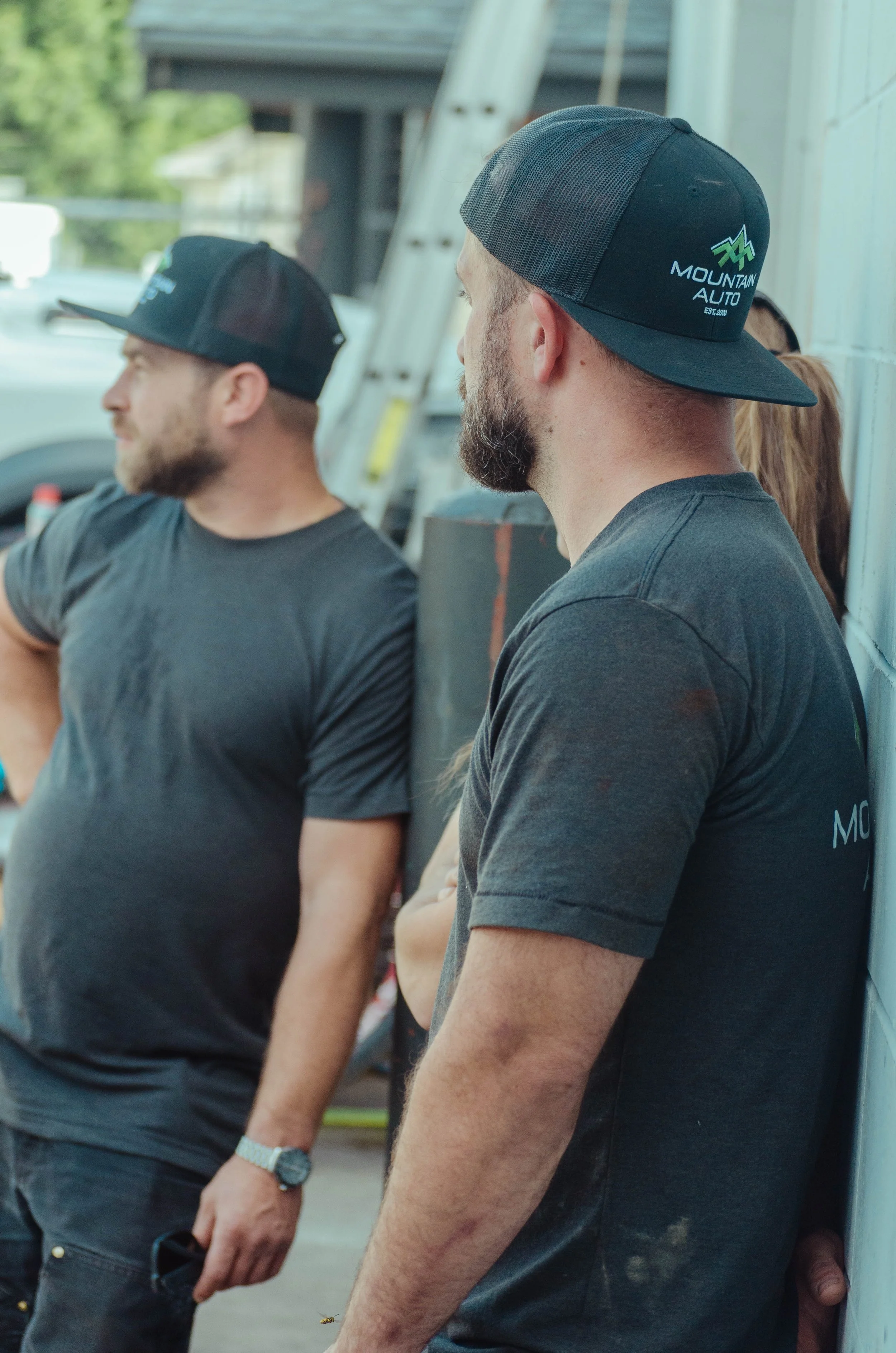 Two men in black t-shirts and black trucker hats standing outdoors, one leaning against a wall with arms crossed, the other looking away, with a ladder and trees in the background.