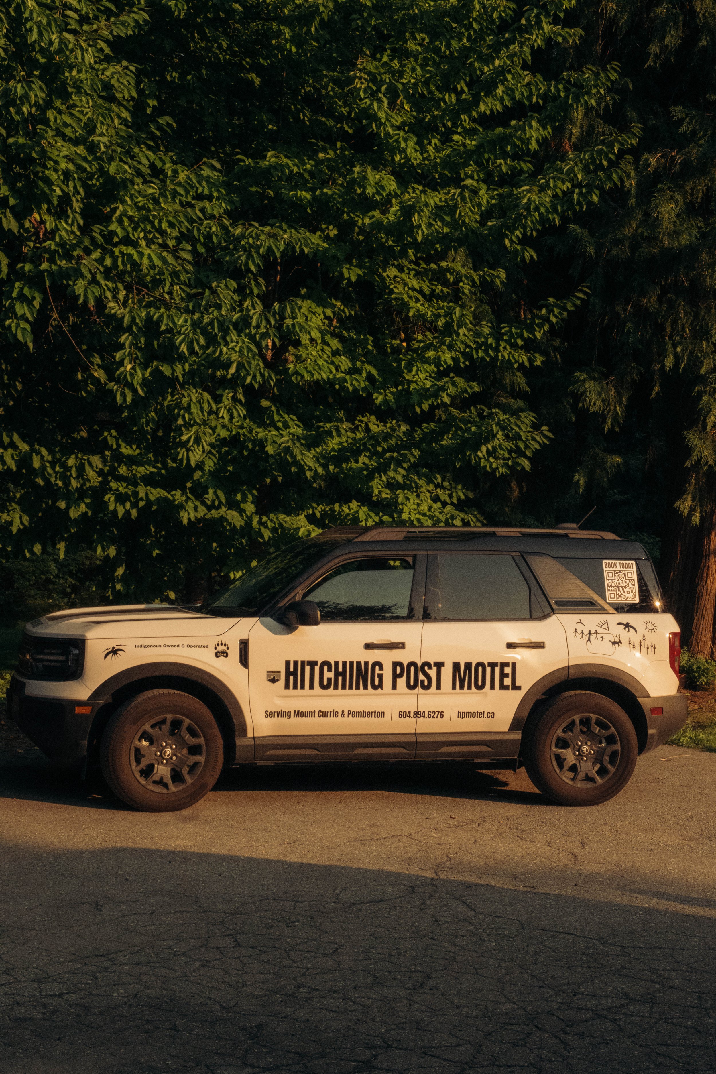 A white SUV with black text and graphics promoting Hitching Post Motel parked on a street in front of green trees at sunset.