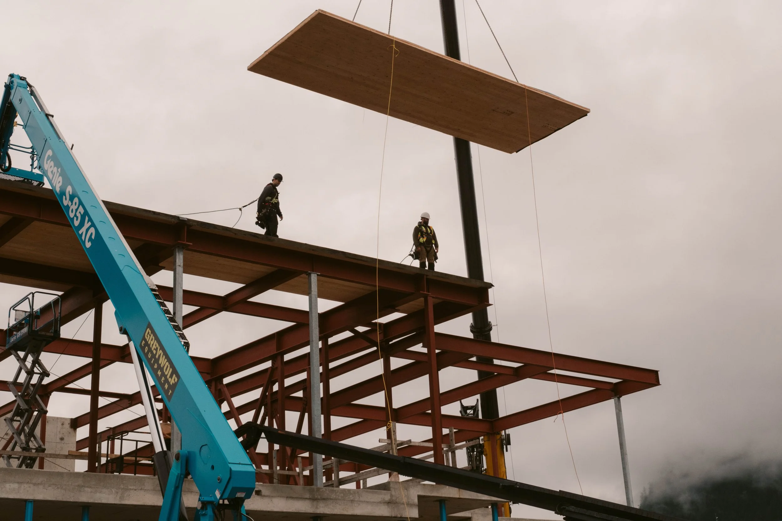 Construction workers on a steel framework building site lifting a large wooden panel to the top of a structure using a crane.
