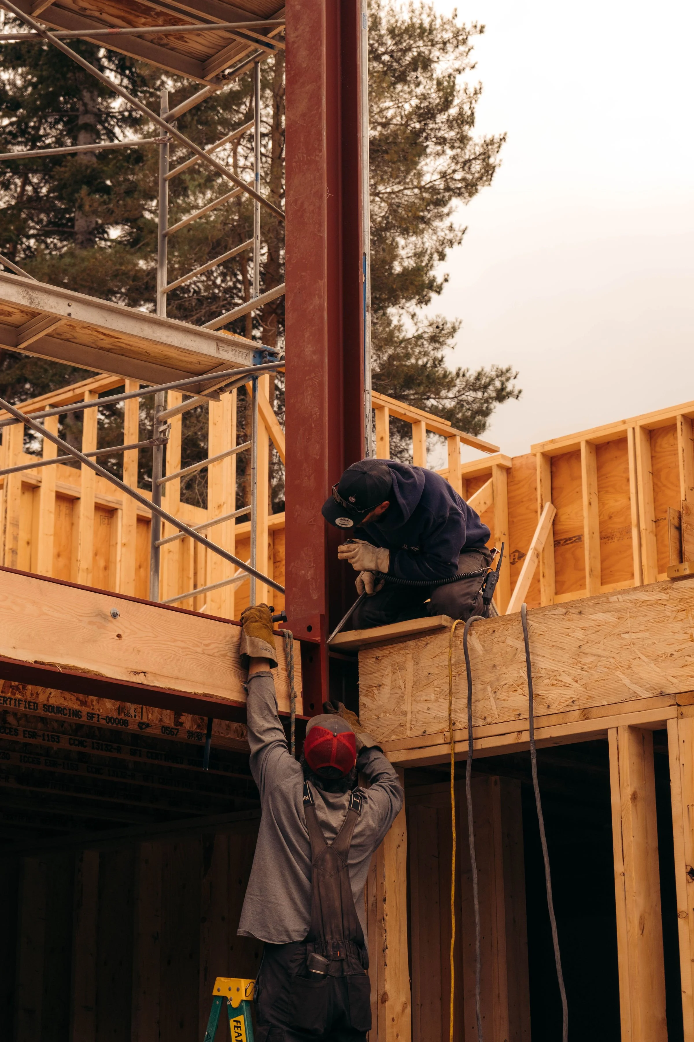 Two construction workers working on a building frame, one standing underneath and holding a beam while the other is seated on the beam securing a steel support, with wooden framing and scaffolding around.