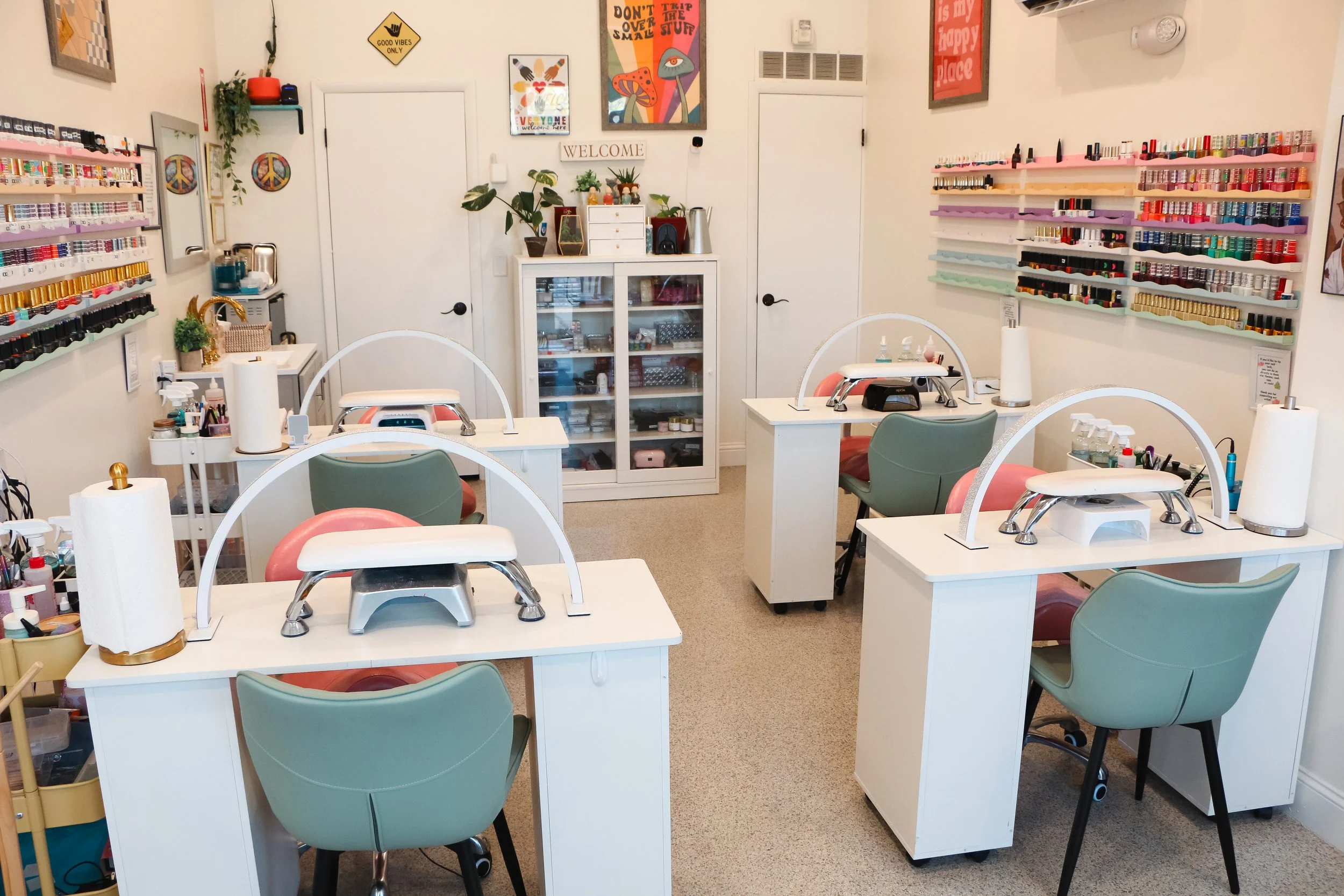 Nail salon with four white tables, each equipped with nail drying lamps and tools. Green and pink chairs sit at each station. Wall-mounted shelves on the left display nail polish bottles. Small cart and storage cabinet are on the left, and artwork de