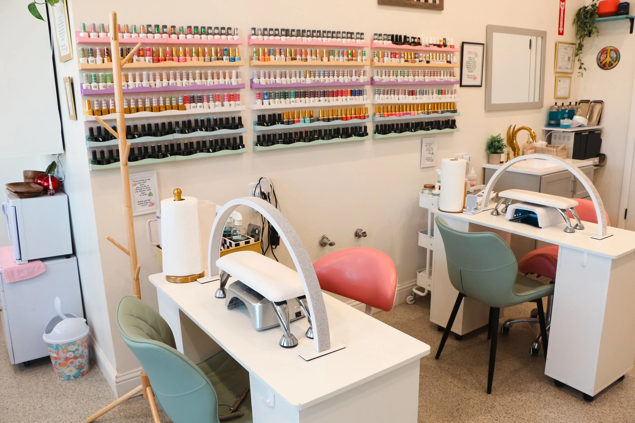 Nail salon with two manicure stations, colorful nail polish bottles on wall-mounted shelves, and various nail care tools and decor.