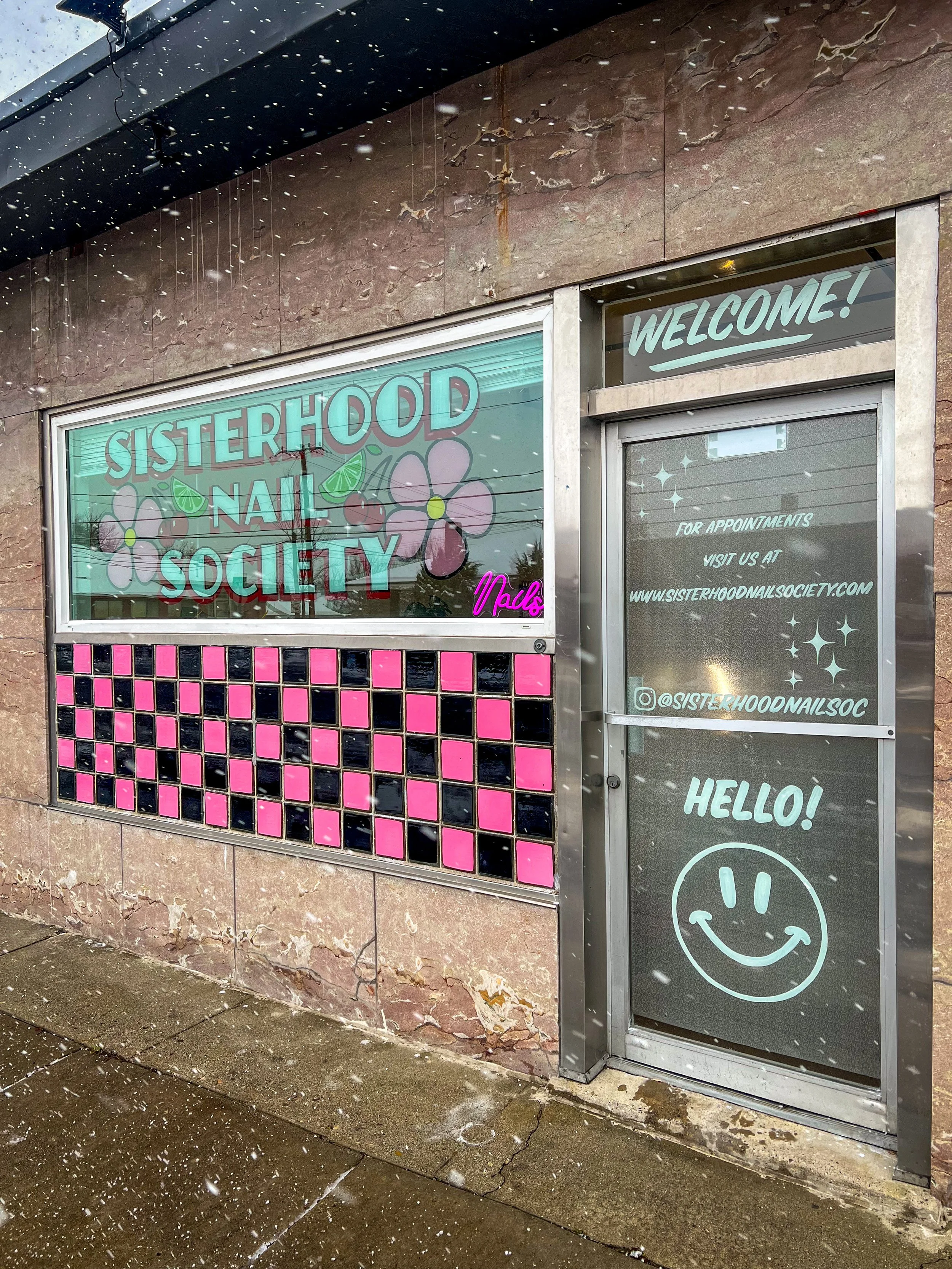 Storefront window with pink and black tile decoration, sign for Sisterhood Nail Society, welcoming message, and a door with a smiley face drawn on it, during snowfall.