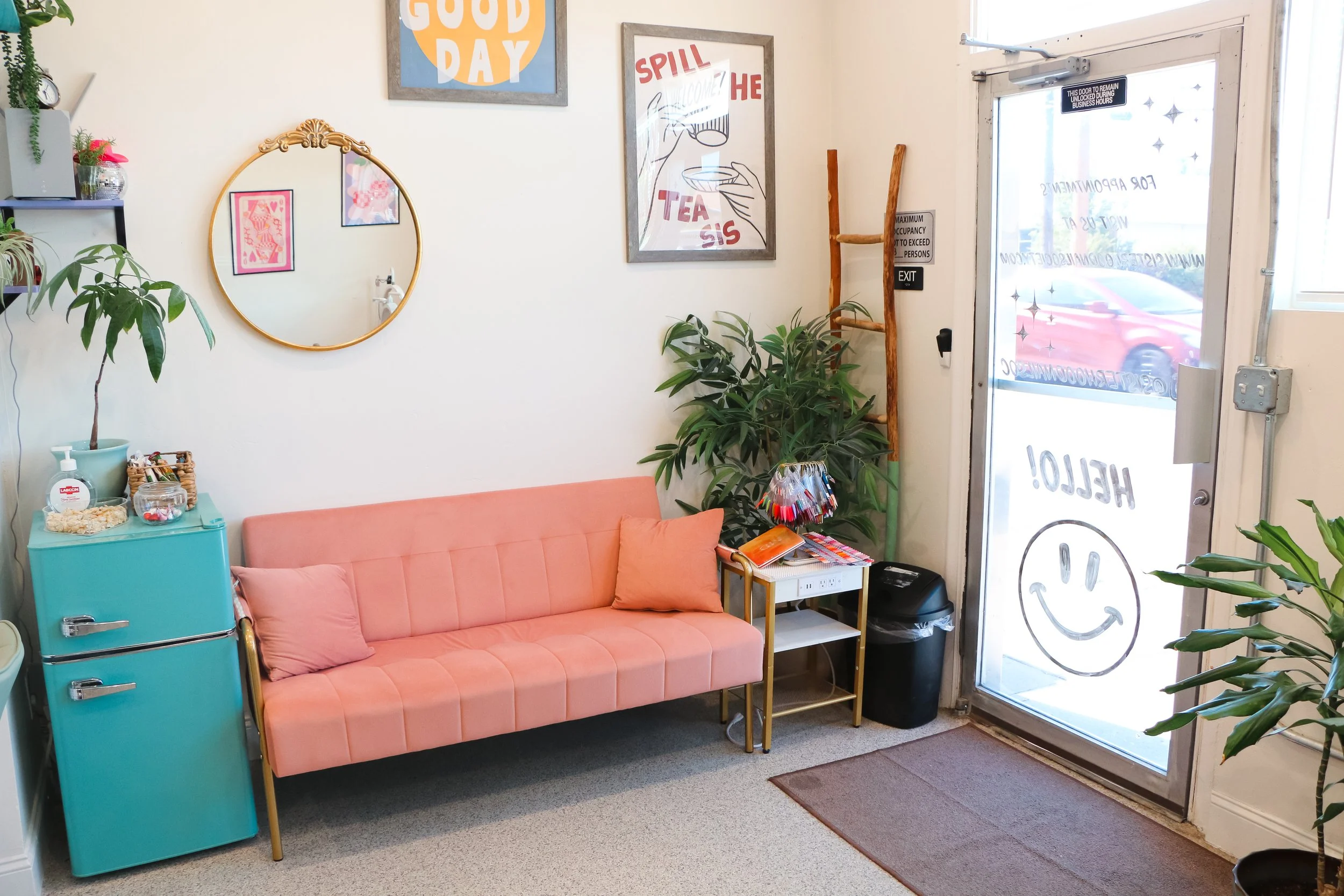 A cozy waiting area with a pink vintage-style sofa, a small teal fridge, a mirror, wall art, an indoor plant, and a glass door with smiley face and welcome message written on it.