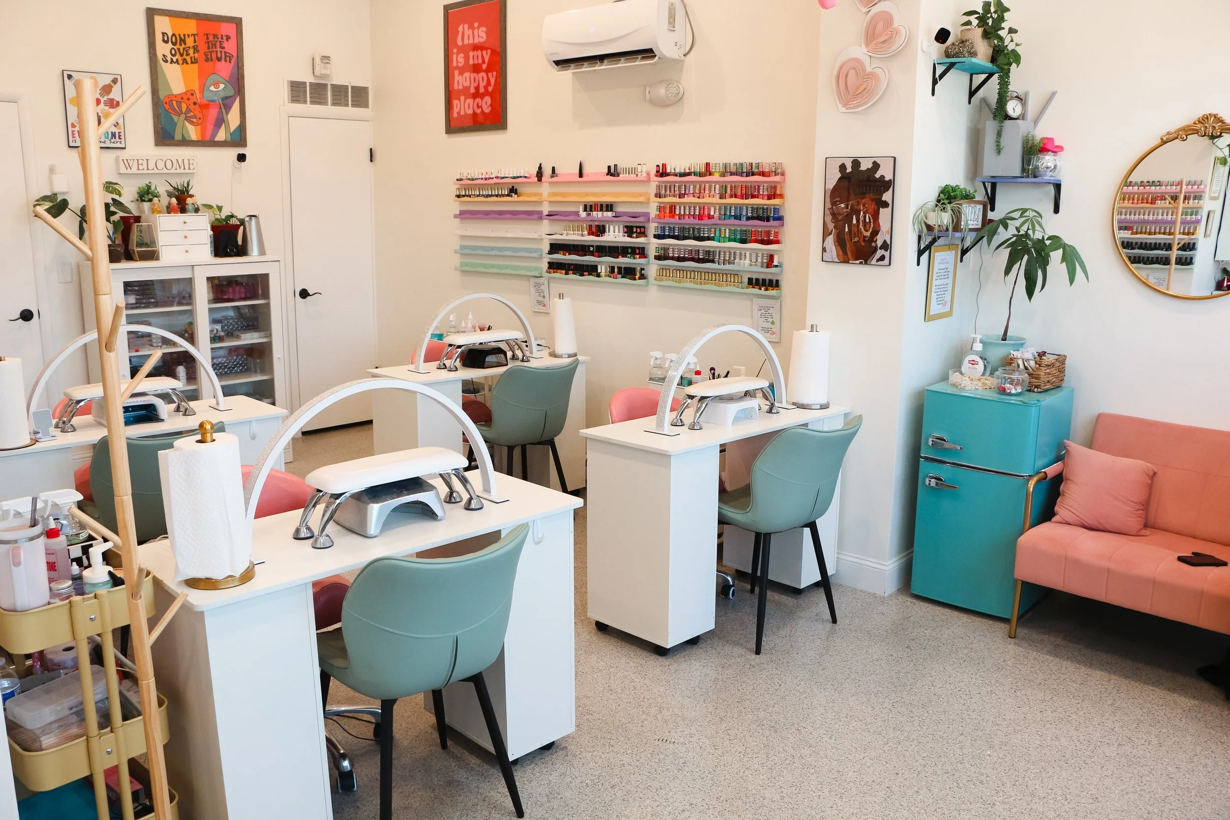 A cozy nail salon with three manicure stations, nail polish racks on the wall, a pink sofa, and decorative wall art in bright, pastel colors.