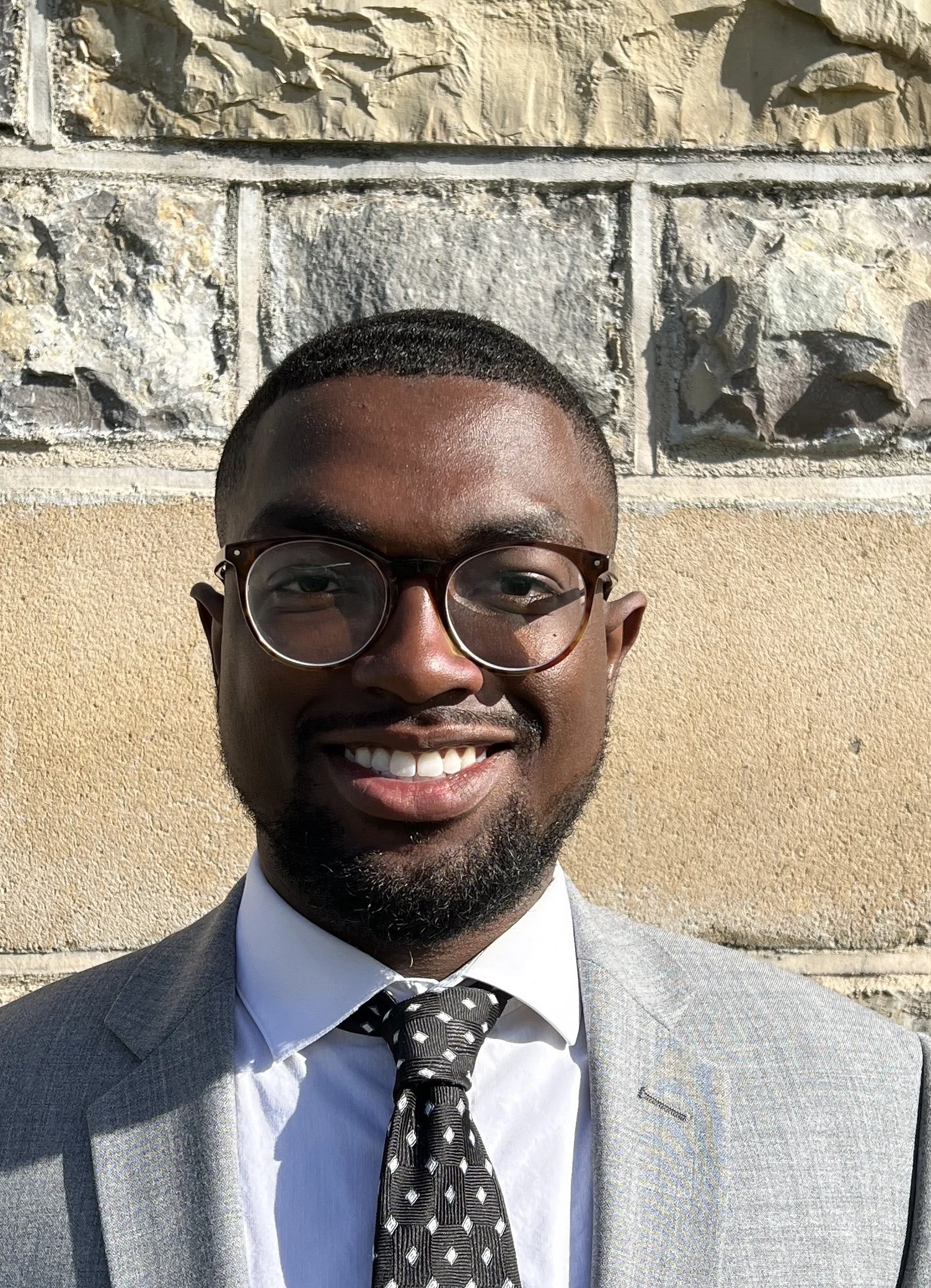A smiling man wearing glasses, a light grey suit, a white shirt, and a black tie with white pattern, standing outside in sunlight against a stone wall background.