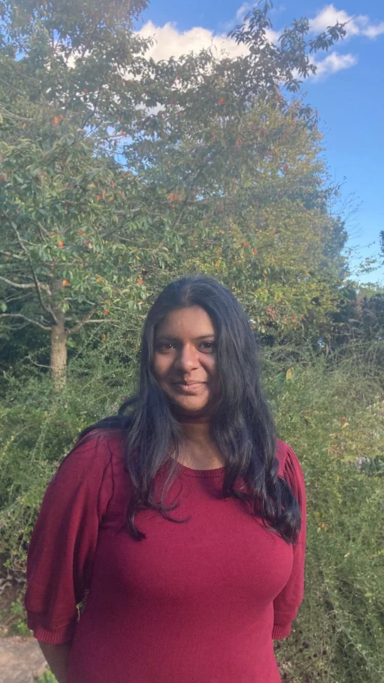 A woman with long black hair wearing a red shirt, standing outdoors with trees and a blue sky in the background.