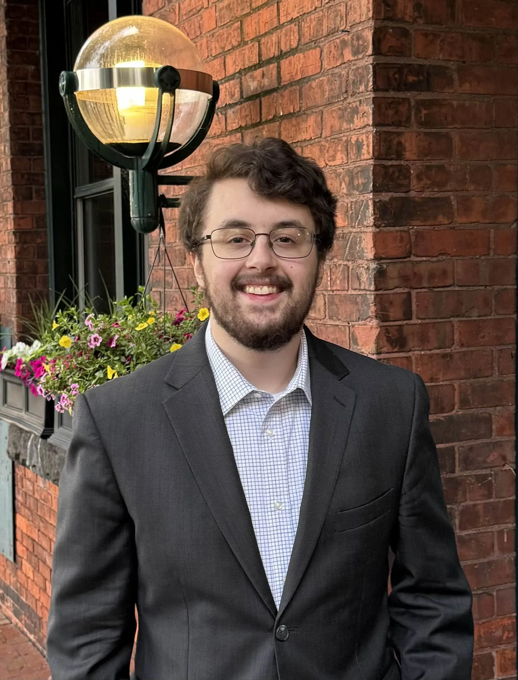 A young man with glasses, brown curly hair, and a beard is standing outdoors at night in front of a brick building. He is dressed in a dark suit jacket over a white checked shirt. Behind him, there is a decorative outdoor lamp and a window box filled with colorful flowers.