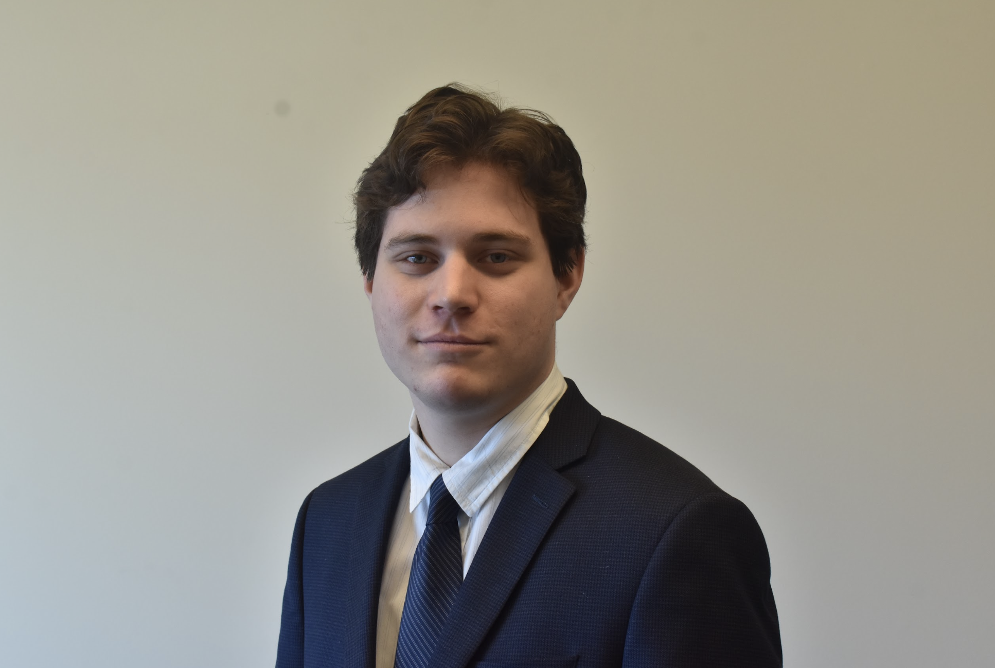 Young man in a dark suit with a striped tie and a white dress shirt, posing against a plain light-colored wall.