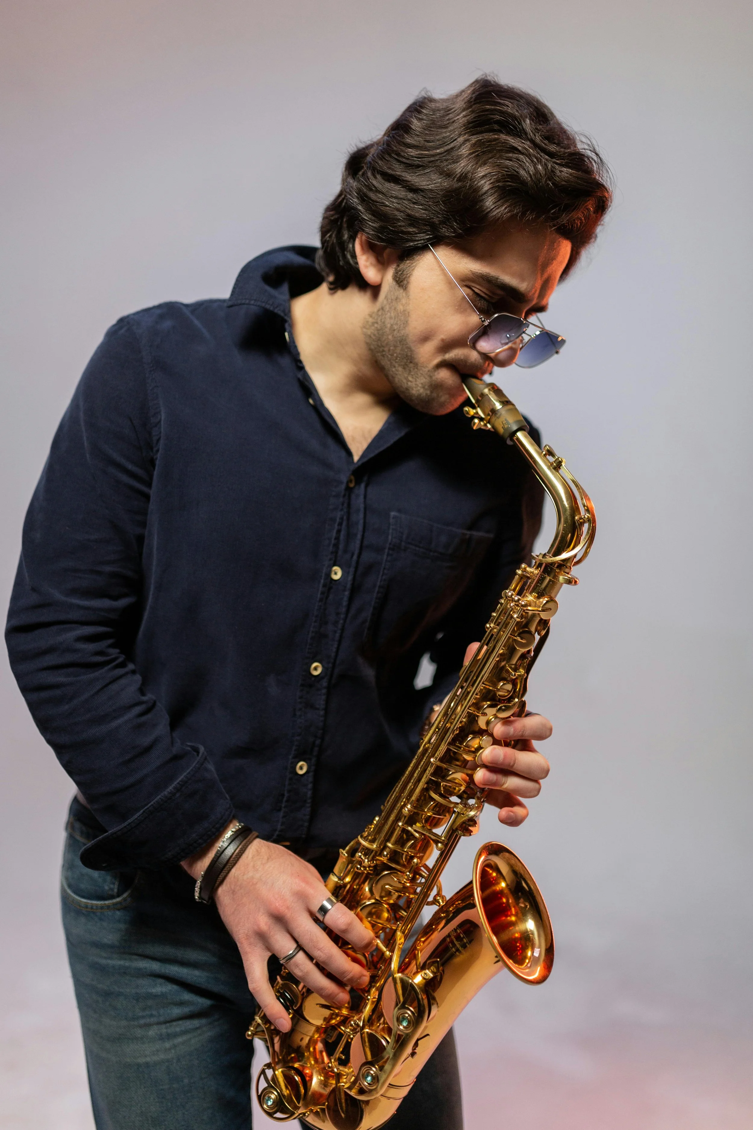 A young man with dark, wavy hair and sunglasses playing a gold-colored saxophone against a plain light gray background.