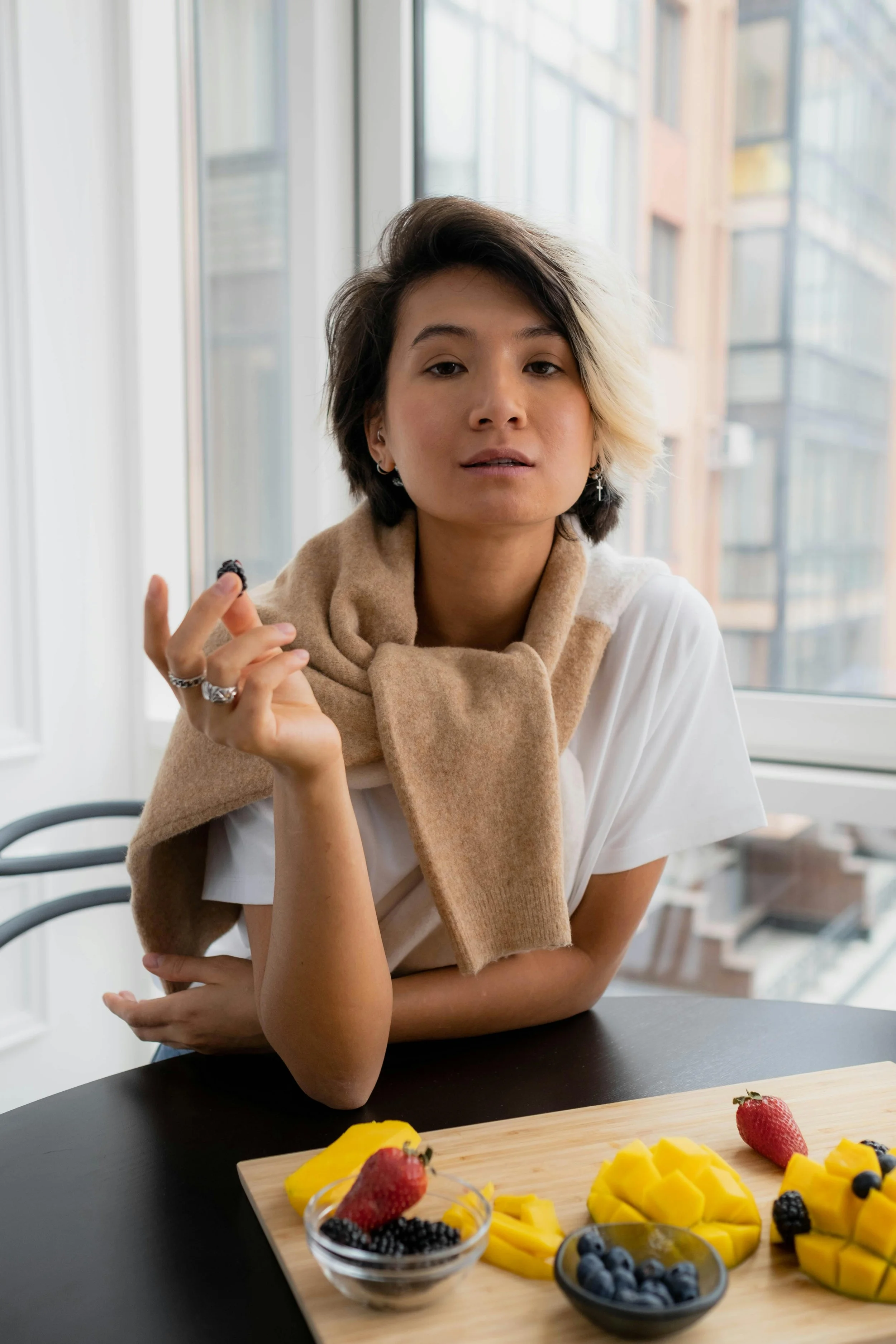 A woman with short hair, wearing earrings and a beige scarf, is sitting at a table near large windows. She is holding a berry in her right hand and looking at the camera. On the table, there is a wooden board with sliced mango, strawberries, and blueberries, and small bowls of berries.