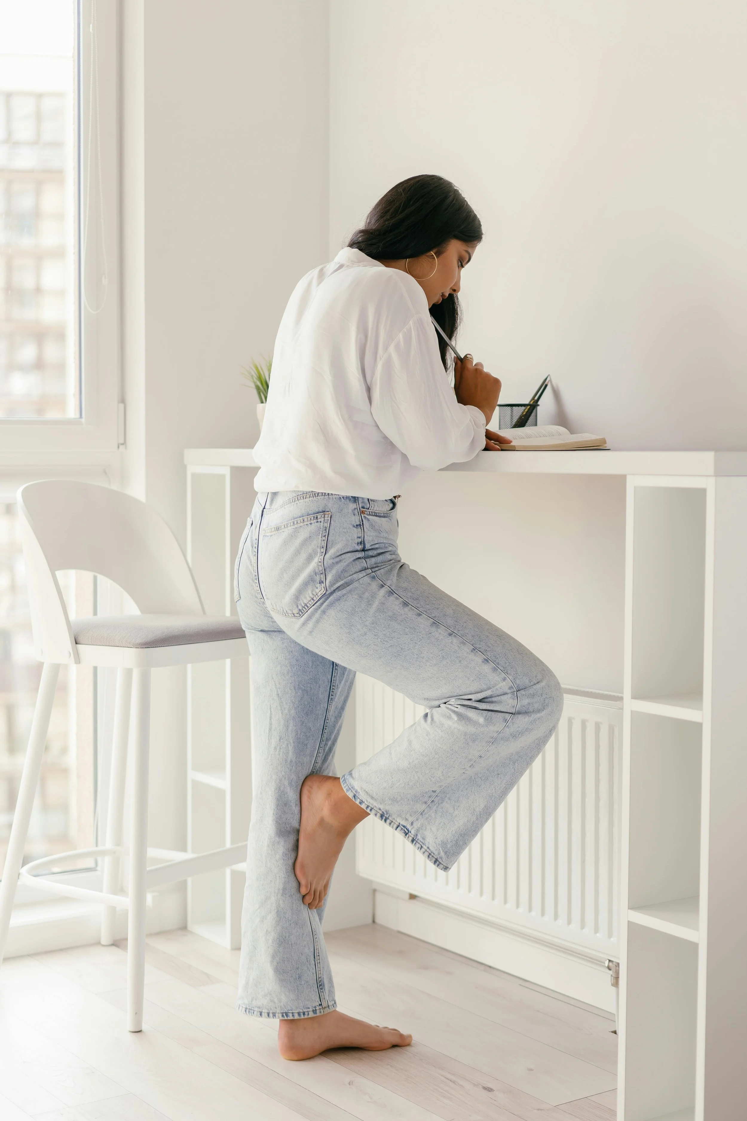A woman standing on one foot, holding a open book on a white desk, with the other foot resting against her leg, in a bright minimalistic room with white walls and a large window.