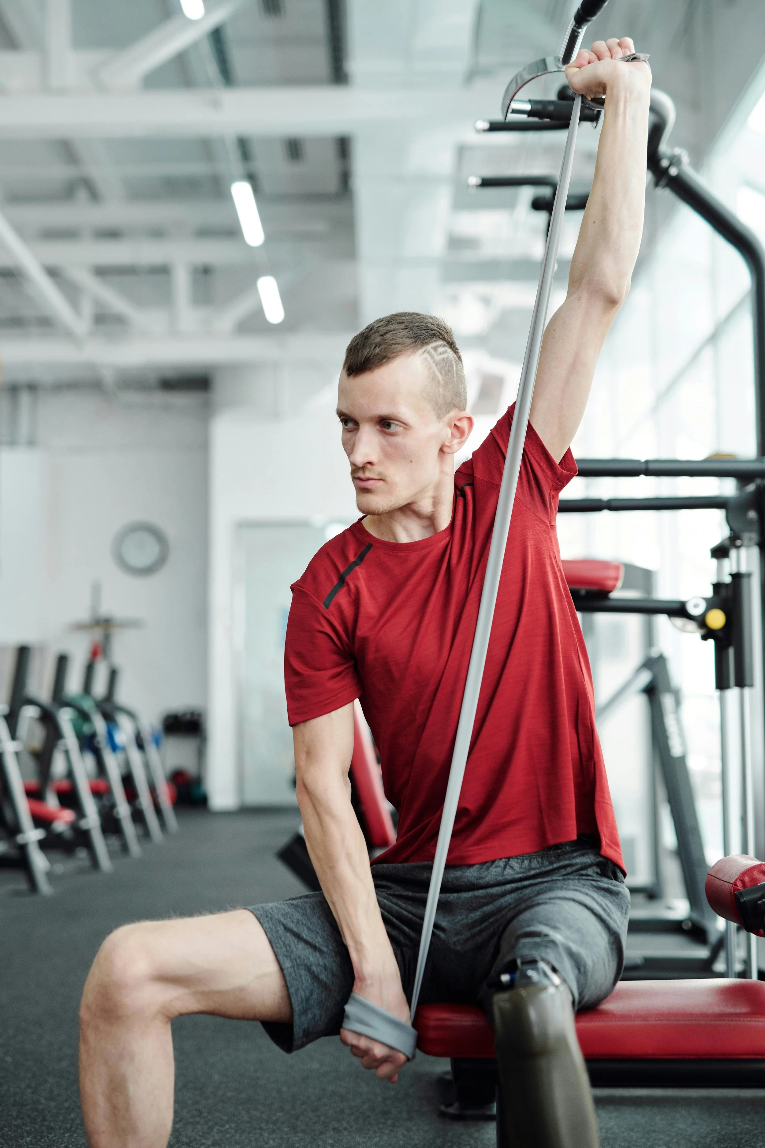 A man is stretching his arm overhead while holding a resistance band, sitting on a workout bench in a gym.