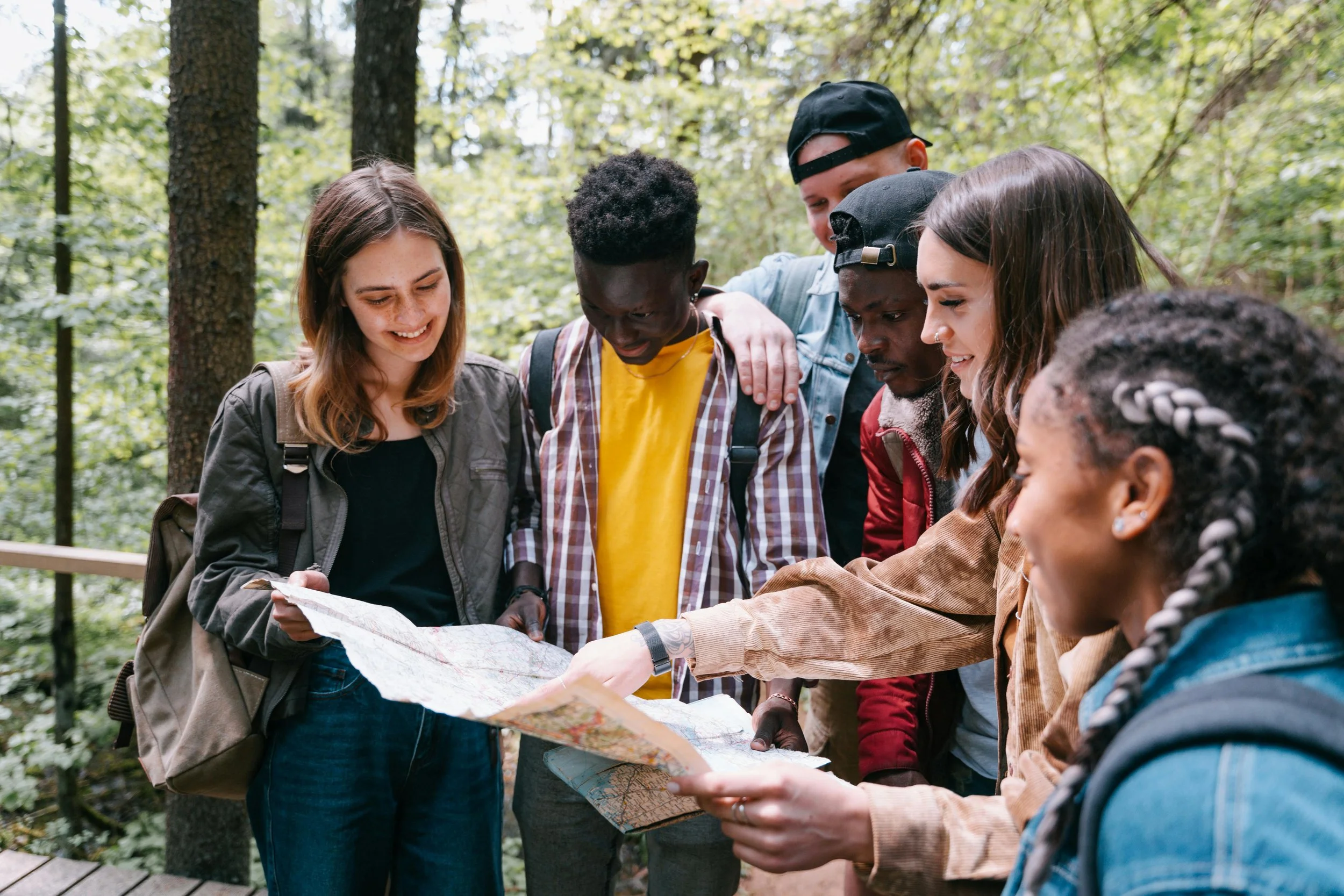 A diverse group of five young people stands closely together outdoors in a wooded area, looking at a map and discussing their location.