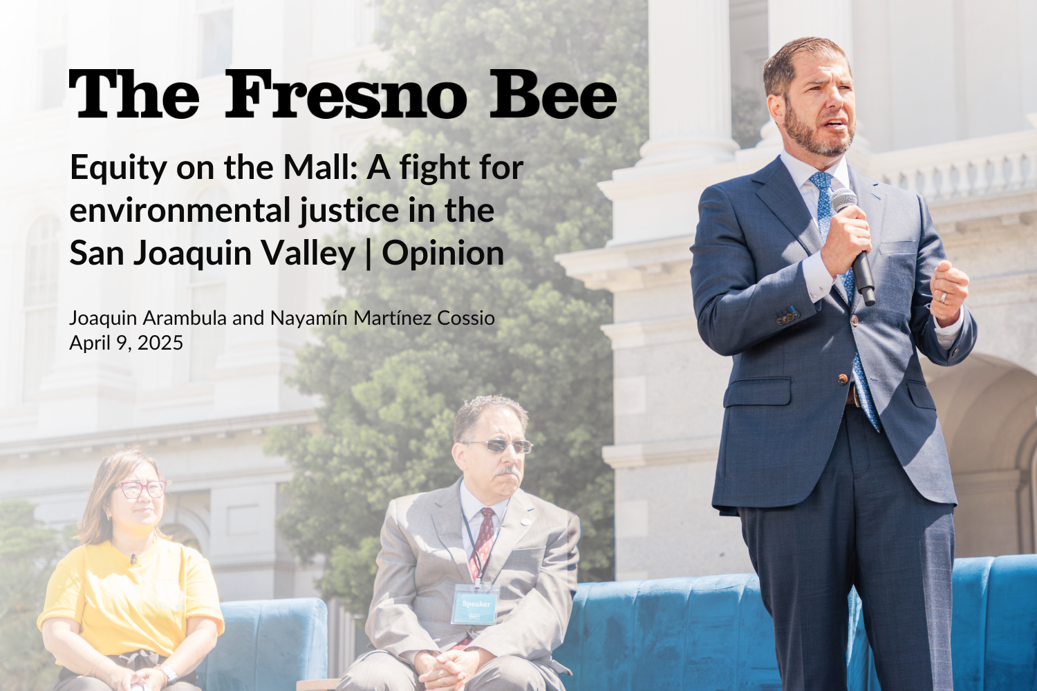 A man in a blue suit speaking into a microphone at an outdoor event in front of a government building, with two seated individuals and a tree in the background.
