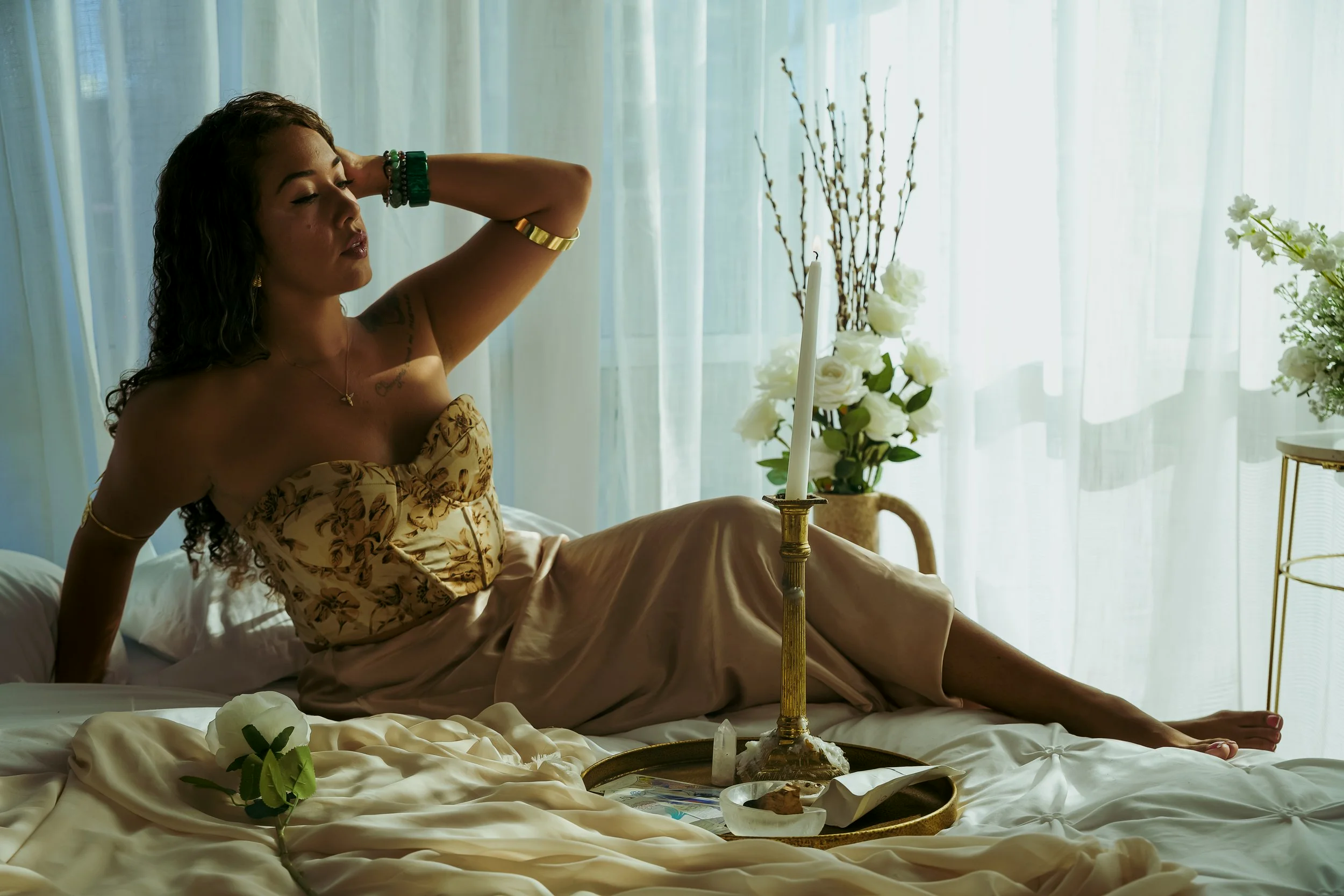 A goddess sitting on a bed in a sunlit room, dressed in elegant clothing, with her eyes closed and one arm resting behind her head. There are flowers and a candle on a tray in front of her. Sanctuary space