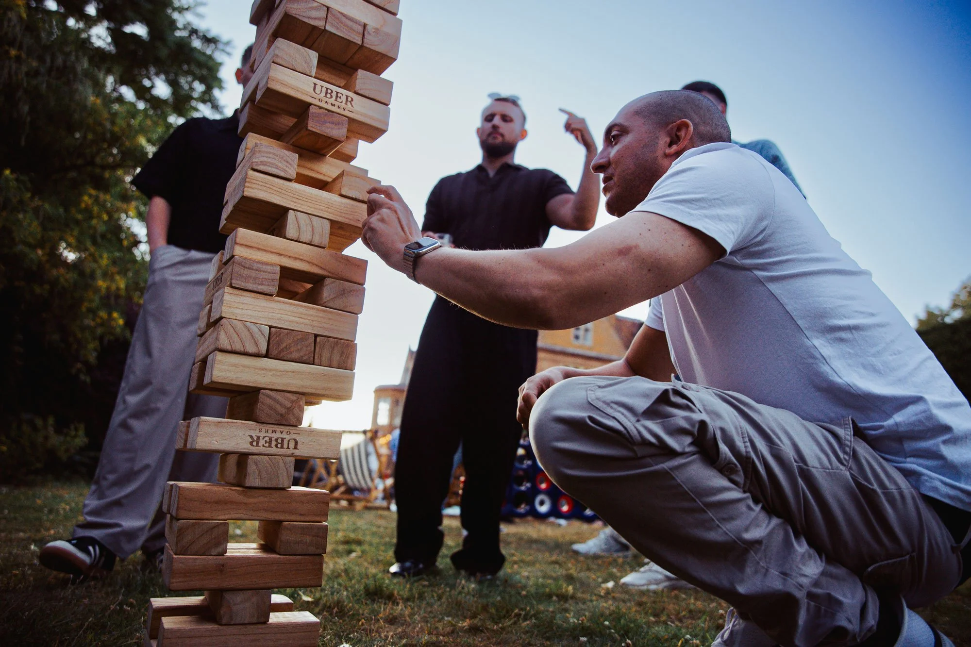Een man concentreert zich vol spanning terwijl hij voorzichtig een houten blok uit een gigantische Jenga-toren trekt op een grasveld, terwijl collega's op de achtergrond toekijken. Dynamische actiefoto op een zomerevenement door SteefClicks.