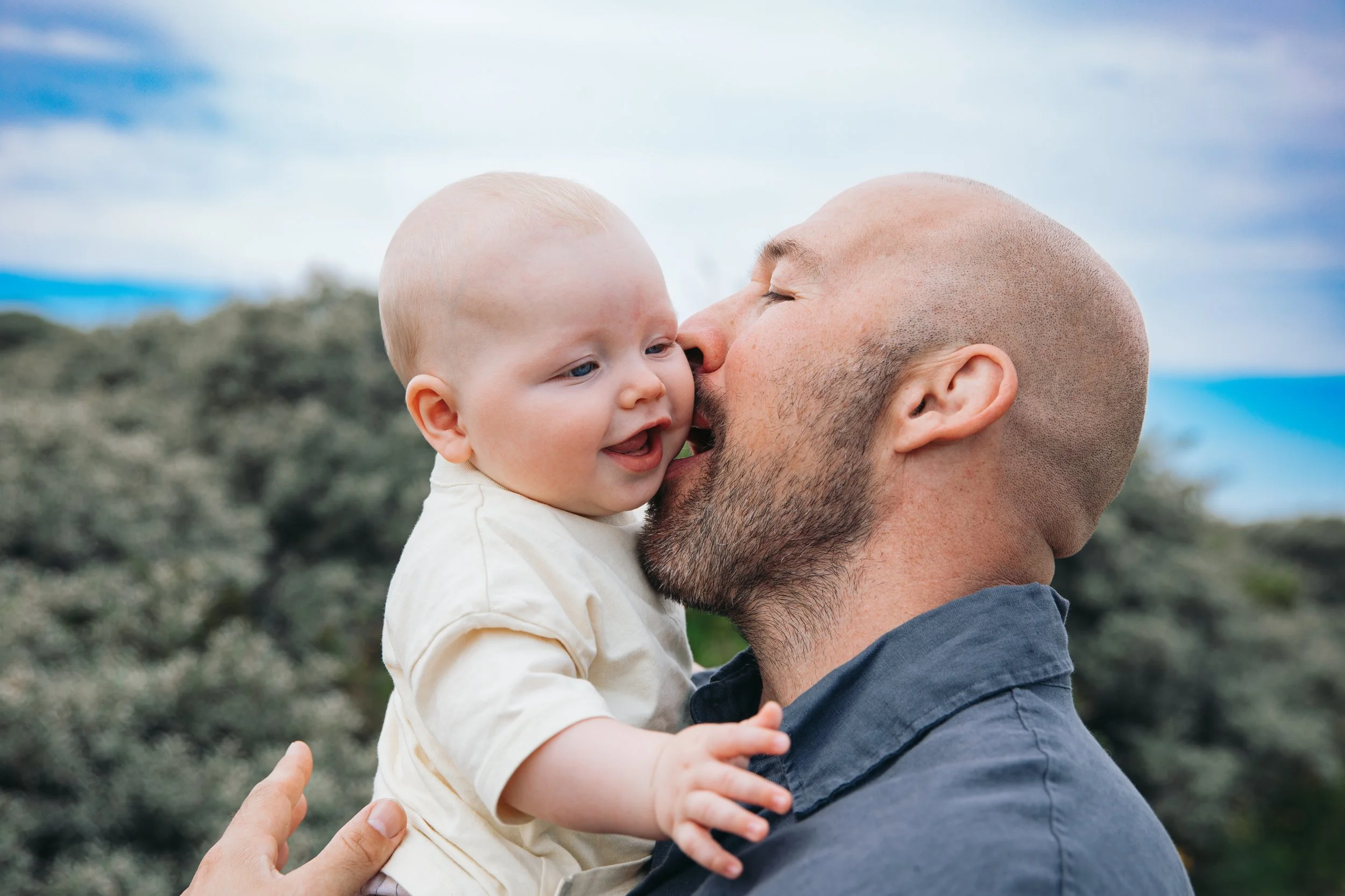 Een man houdt een glimlachende baby in de buitenlucht, beiden met kaal hoofd, tegen een landschap met een blauwe lucht en wolken.