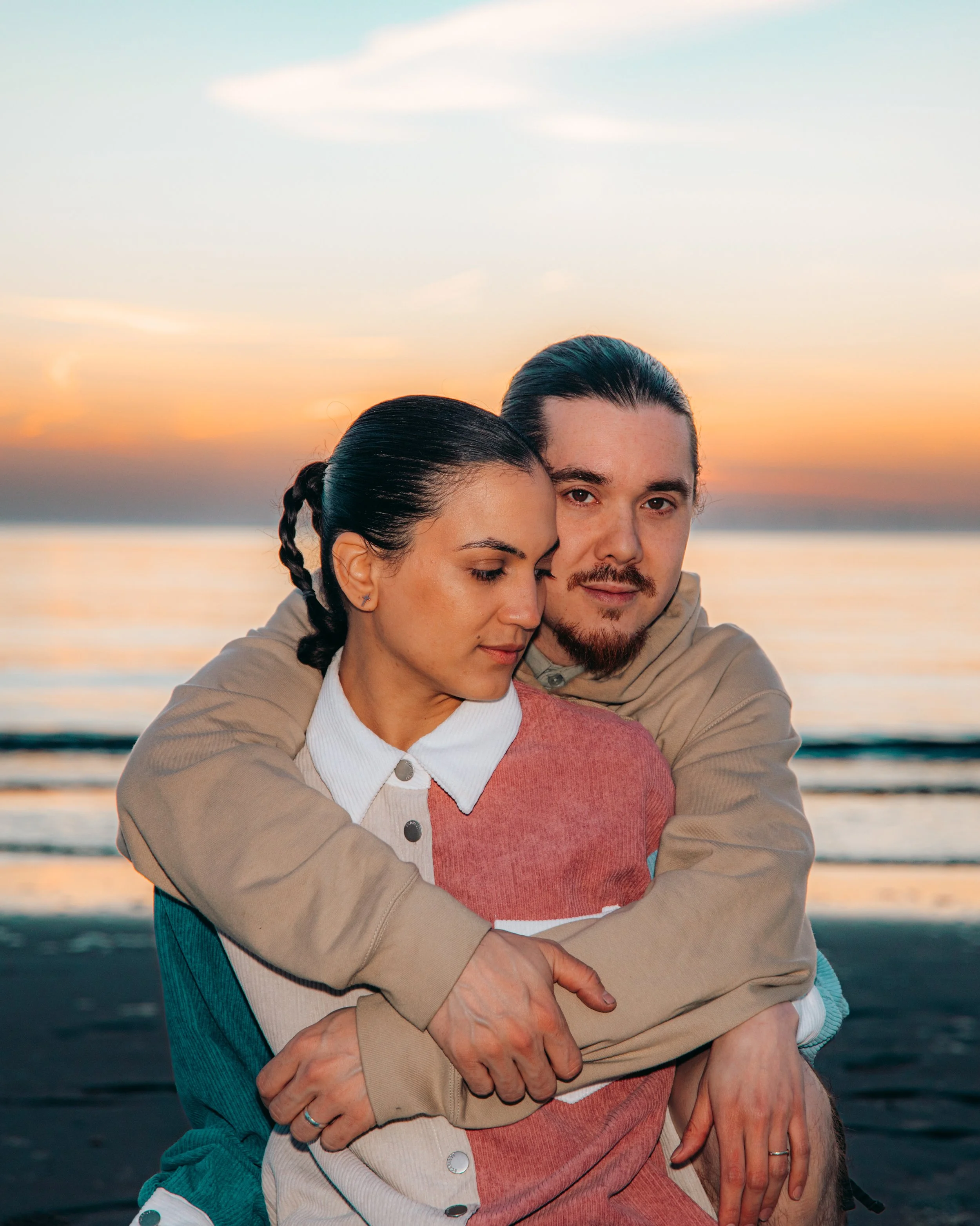 Een man en een vrouw omhelzen elkaar op een strand bij zonsondergang, met de oceaan op de achtergrond.