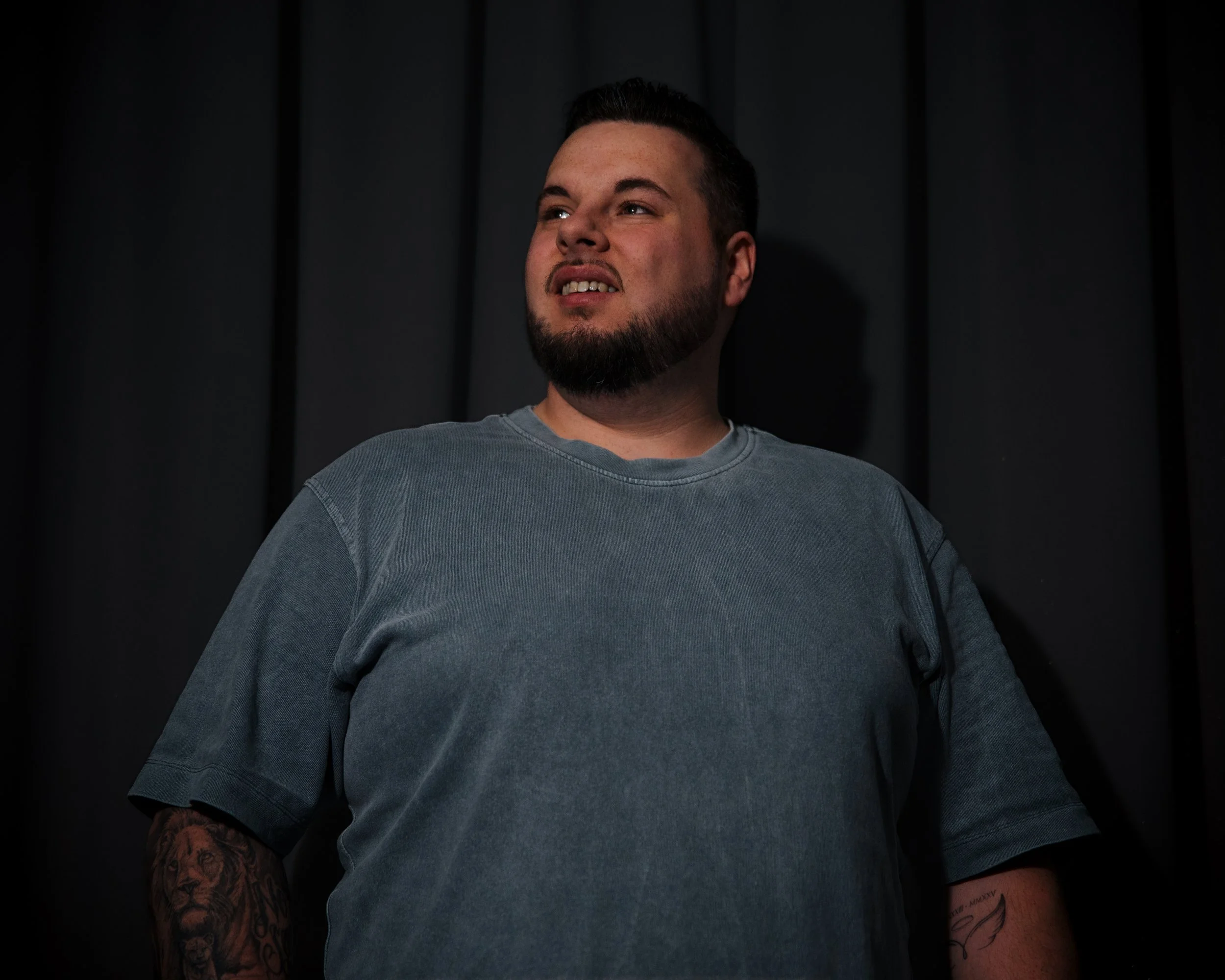 Man met een baard en tattoos op de armen, gekleed in een grijze t-shirt, staat voor een zwarte achtergrond.