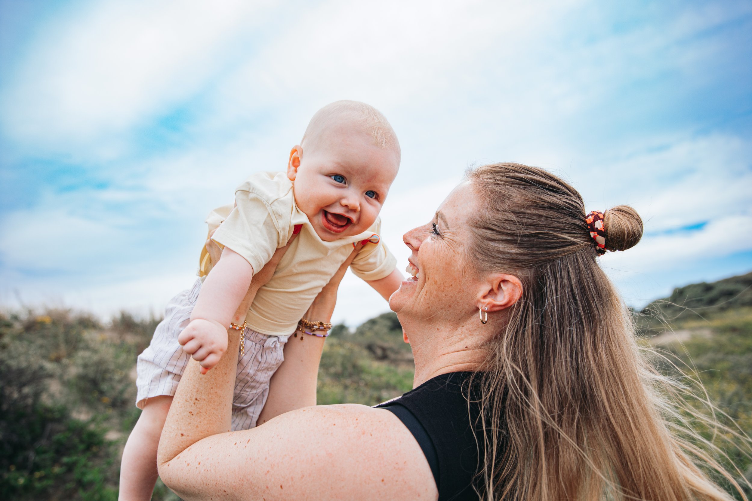 Een vrouw houdt een lachende baby omhoog in een open veld tegen een blauwe lucht met wolken.