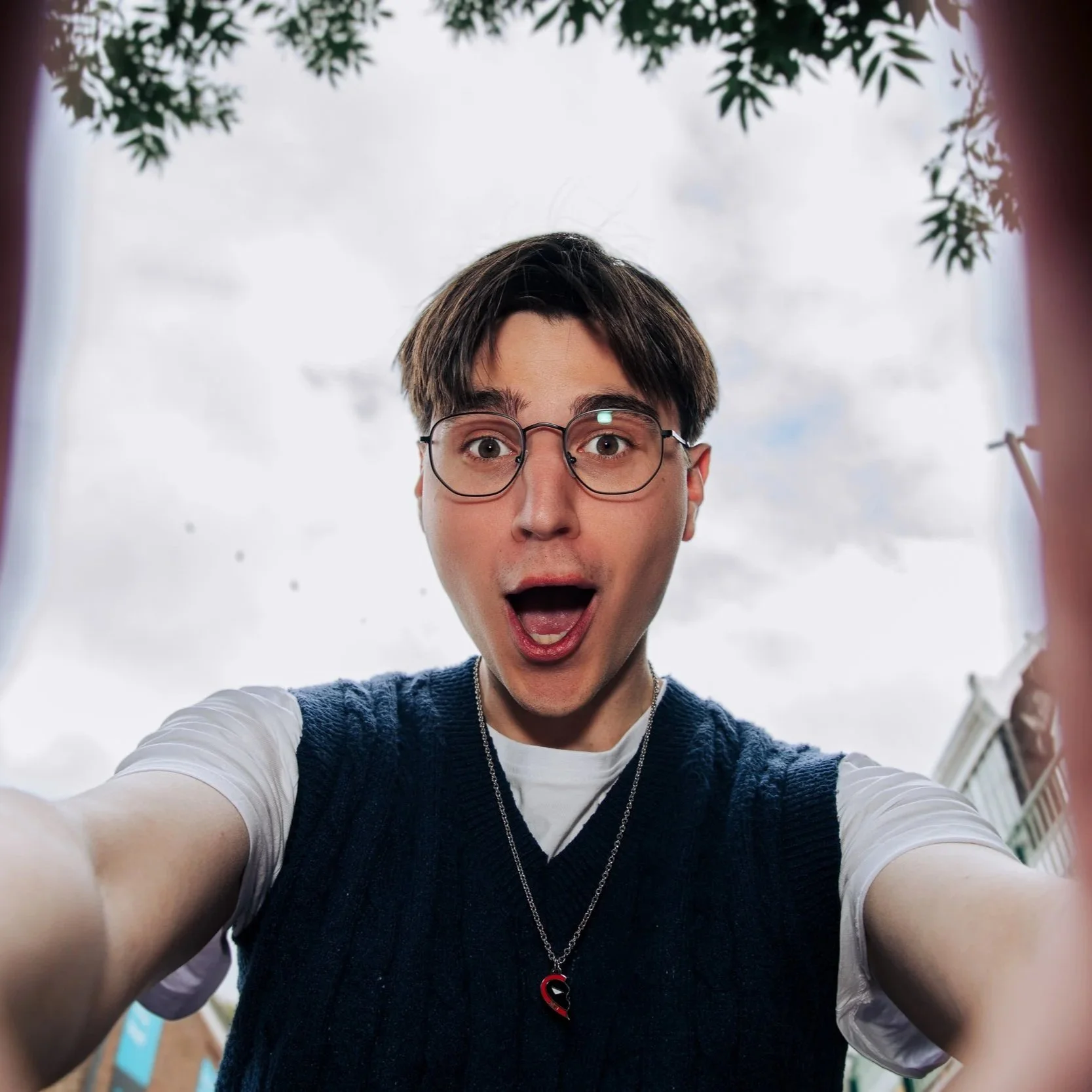 Close-up van een jonge man met bruin haar en bril die een selfie neemt, met een blauwe jas en een getailleerde witte T-shirt, onder een bewolkte hemel.