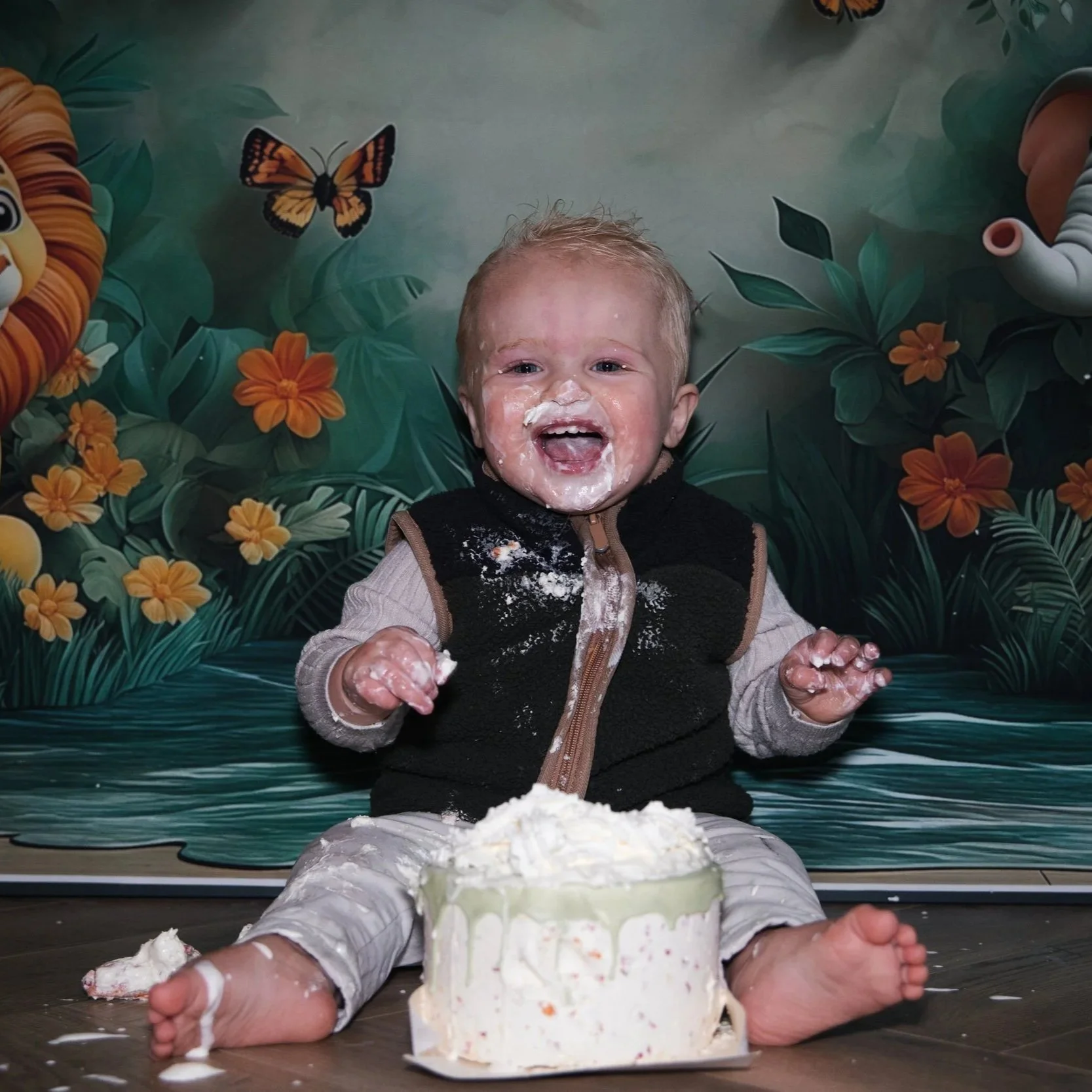 Een blije baby boy lacht breeduit met frosting op zijn gezicht tijdens een cakesmash fotoshoot bij SteefClicks. Hij zit voor een jungle-thema achtergrond met een leeuw en olifant, met een taart voor zich. Locatie Leidschendam.