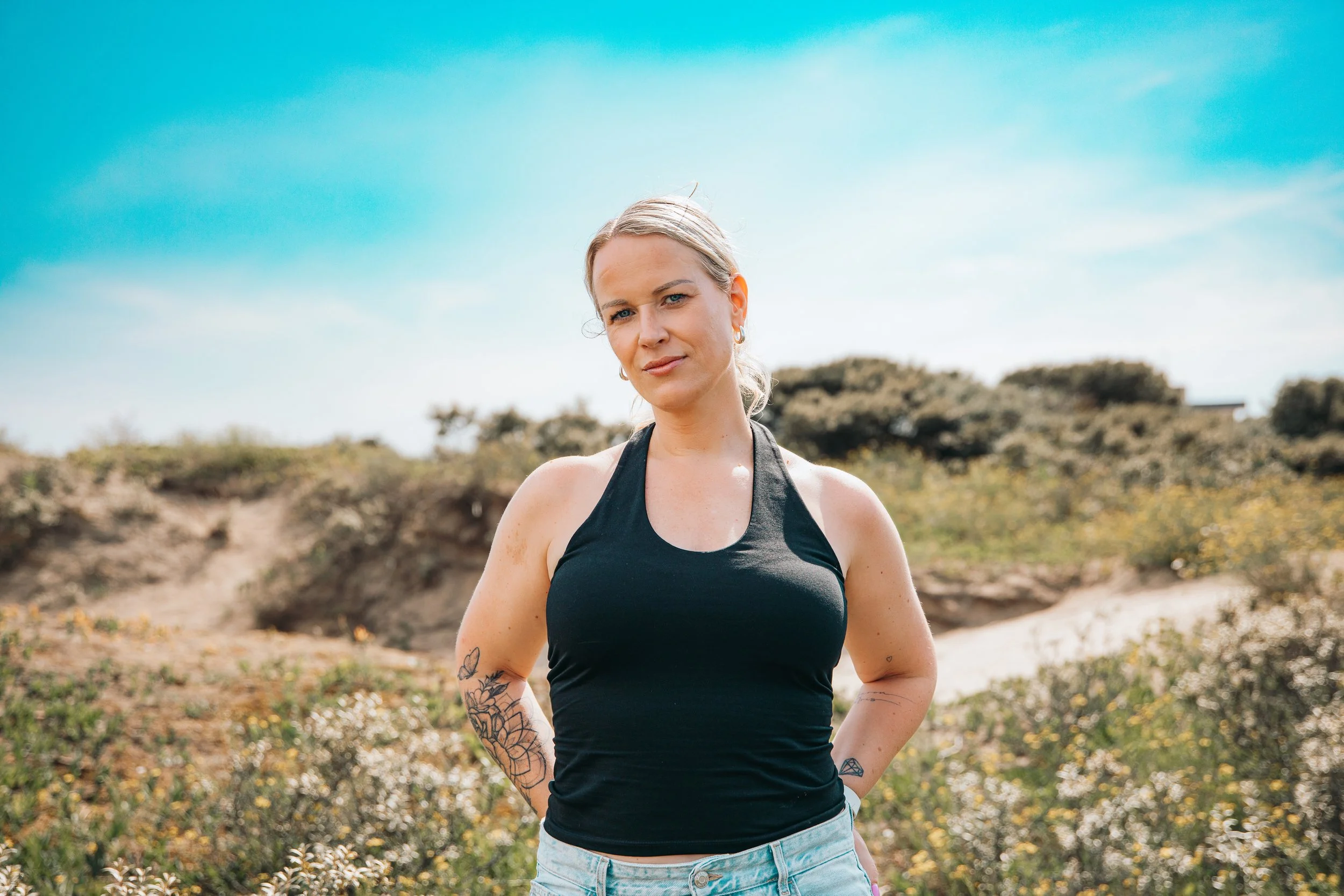 Vrouw met blonde haar, blauwe ogen, sportoutfit en tatoeages poseert buiten onder een blauwe lucht. Foto door SteefClicks / Steef
