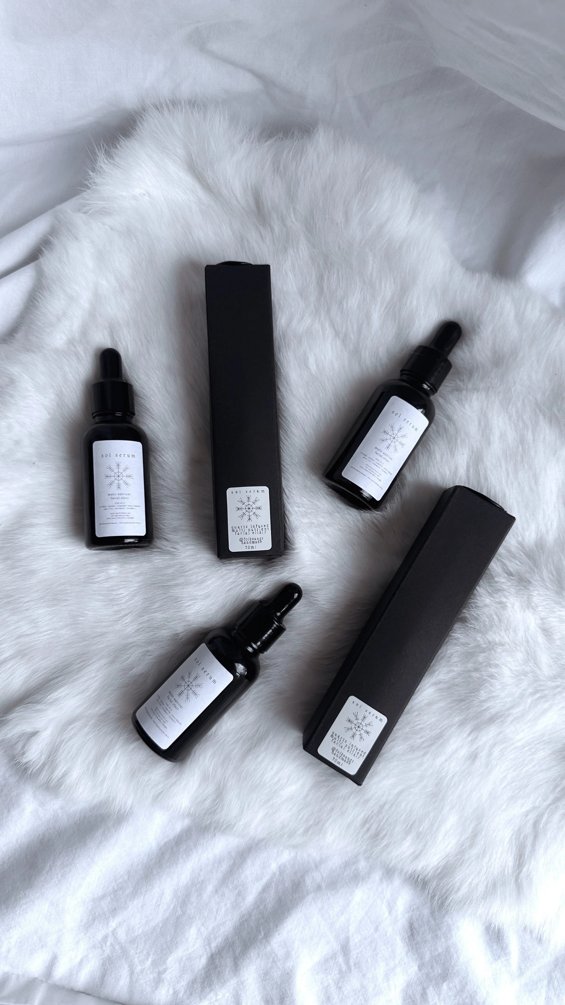Black glass facial serum bottles with white labels featuring the Ægishjálmr, "helm of awe" symbol, displayed on white fur.