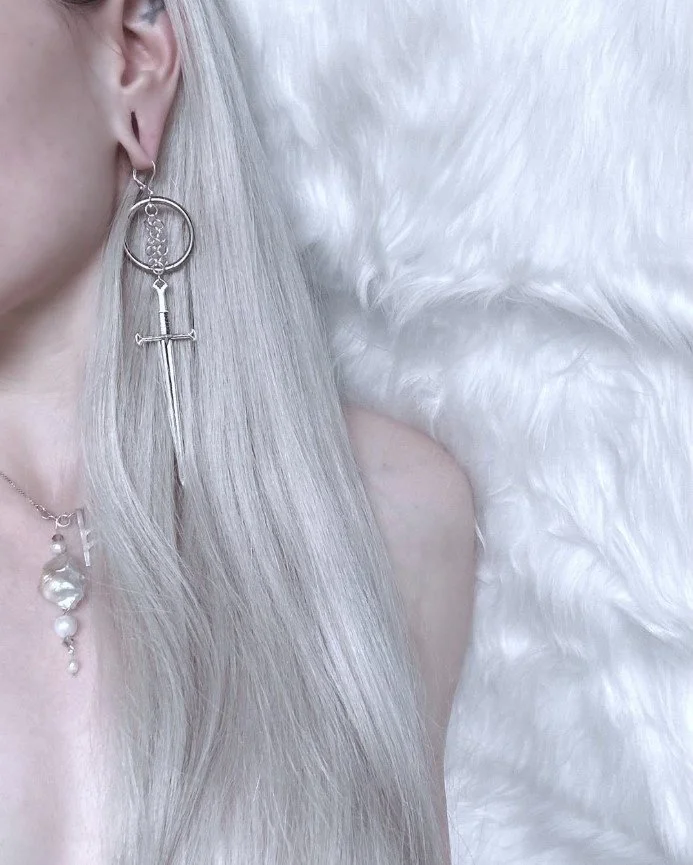 Close-up of a woman with white long hair's side wearing handmade chainmaille sword earrings and a rune and pearl necklace.