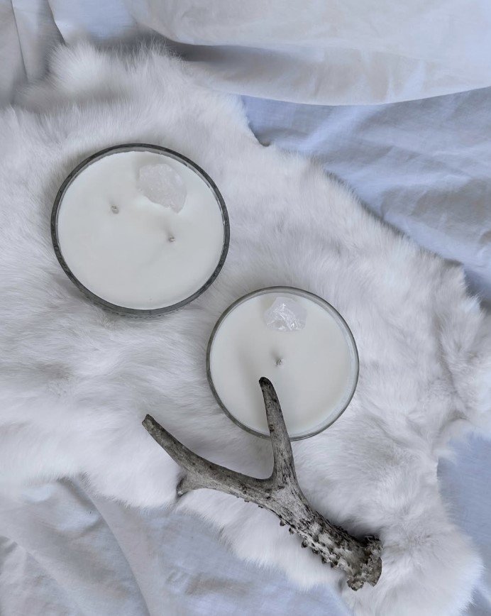 Two white candles in glass holders are placed on a white faux fur surface. An antler is positioned between the candles, creating a decorative arrangement.