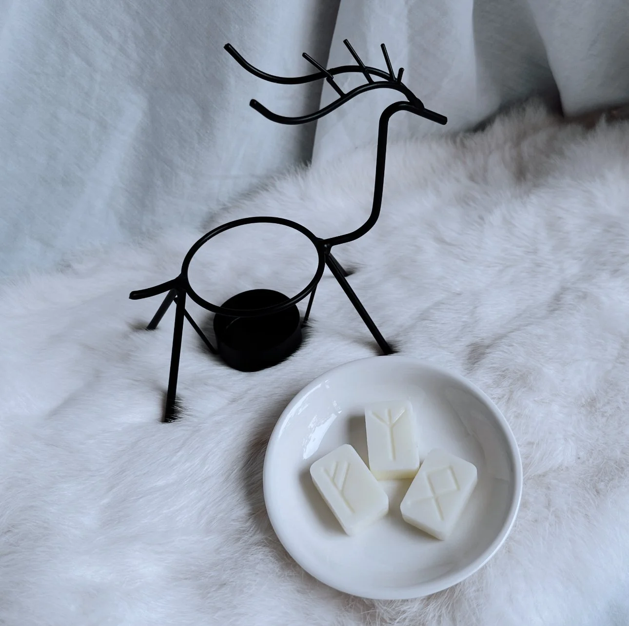 Black reindeer-shaped metal candle holder with a small black candle, and a white ceramic dish with four white wax melt bars engraved with runic symbols, all placed on a white furry rabbit pelt surface.