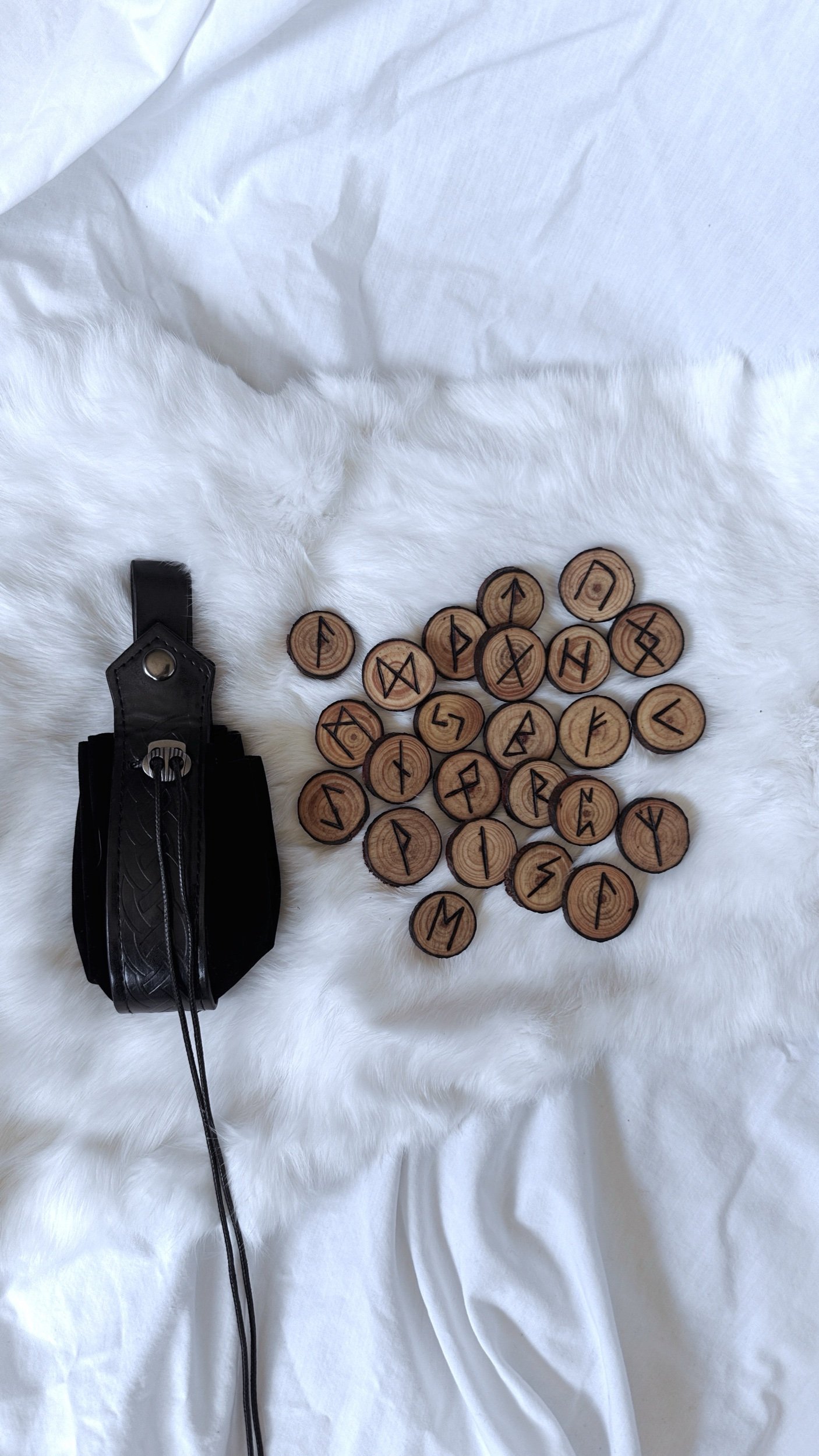 A collection of wooden rune stones with symbols and a black leather strap attached to a smaller black case, all placed on a white, fuzzy fabric background.