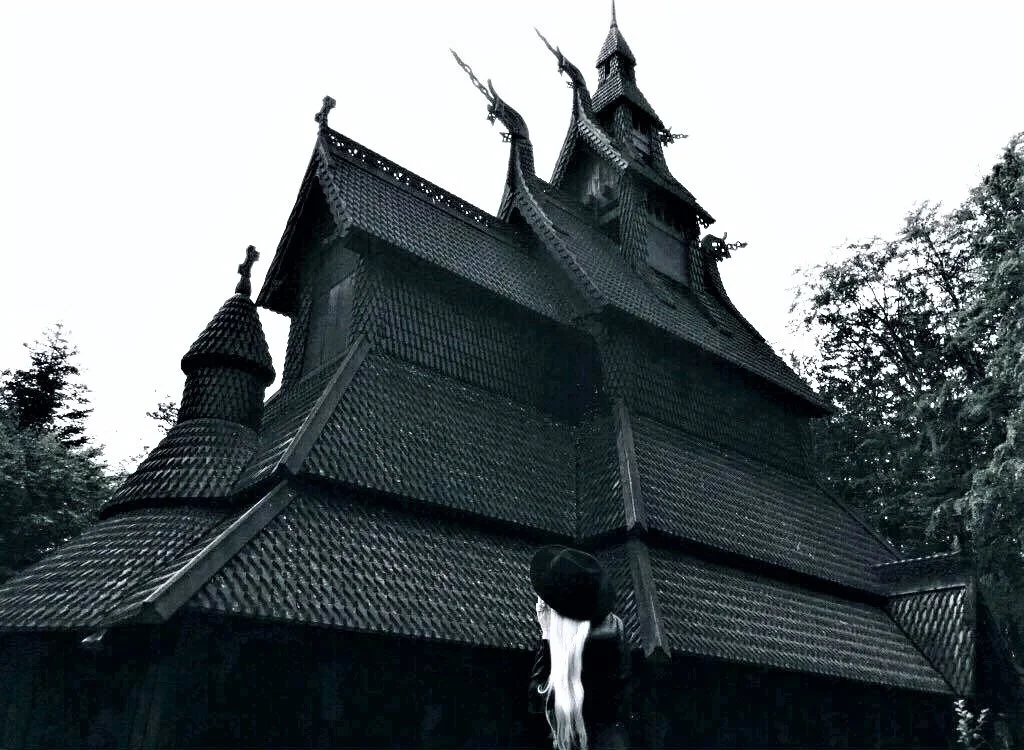 Woman with long white hair standing in front of a Norwegian Stave church, surrounded by trees in a black and white photograph.