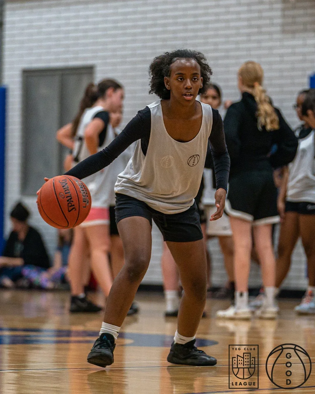 Young girl dribbling a basketball on an indoor court with other players and a coach in the background.