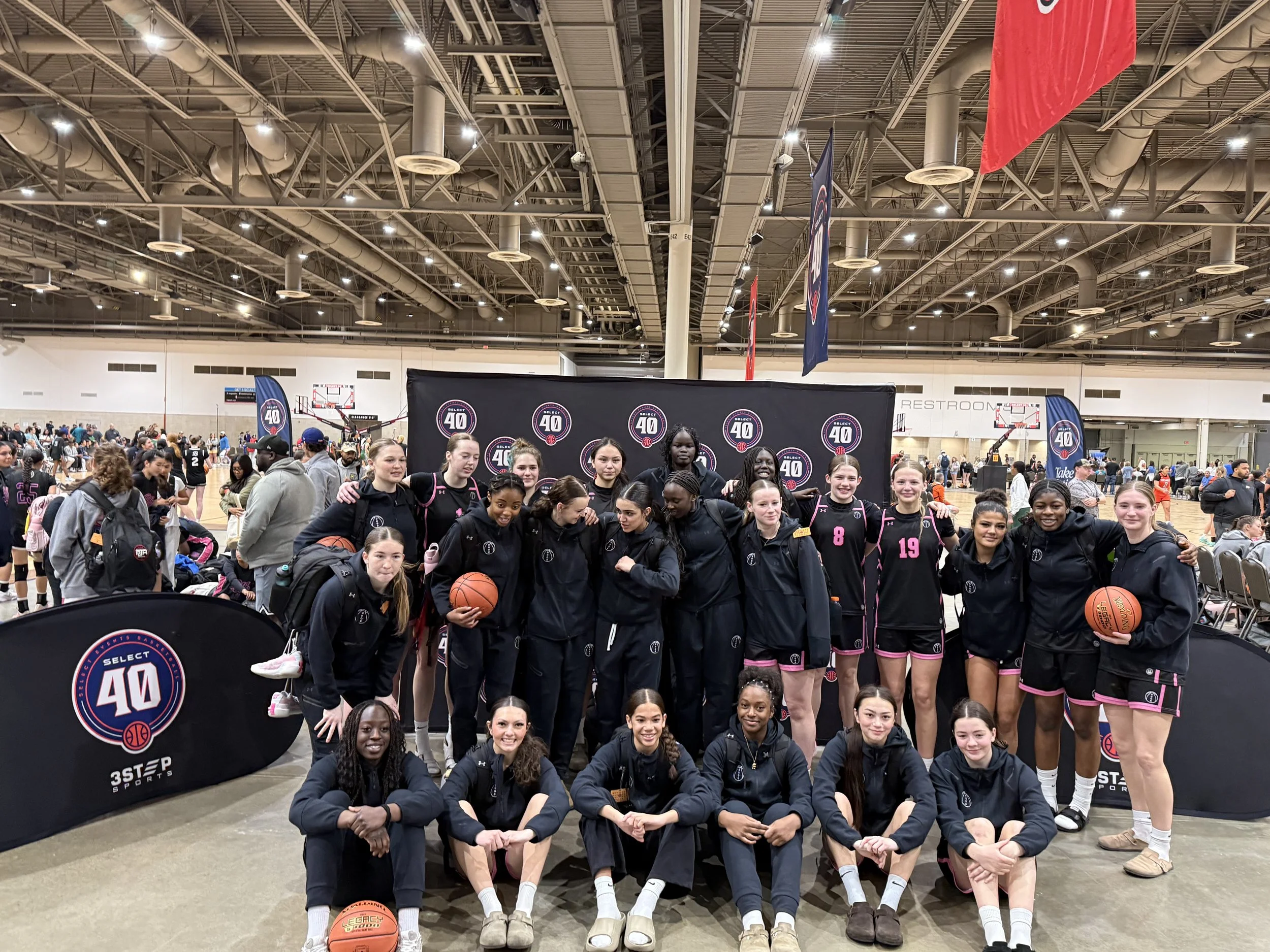 A group of female basketball players in black and white uniforms gathered in a gym during a timeout, some sitting and others standing, listening to their coach.
