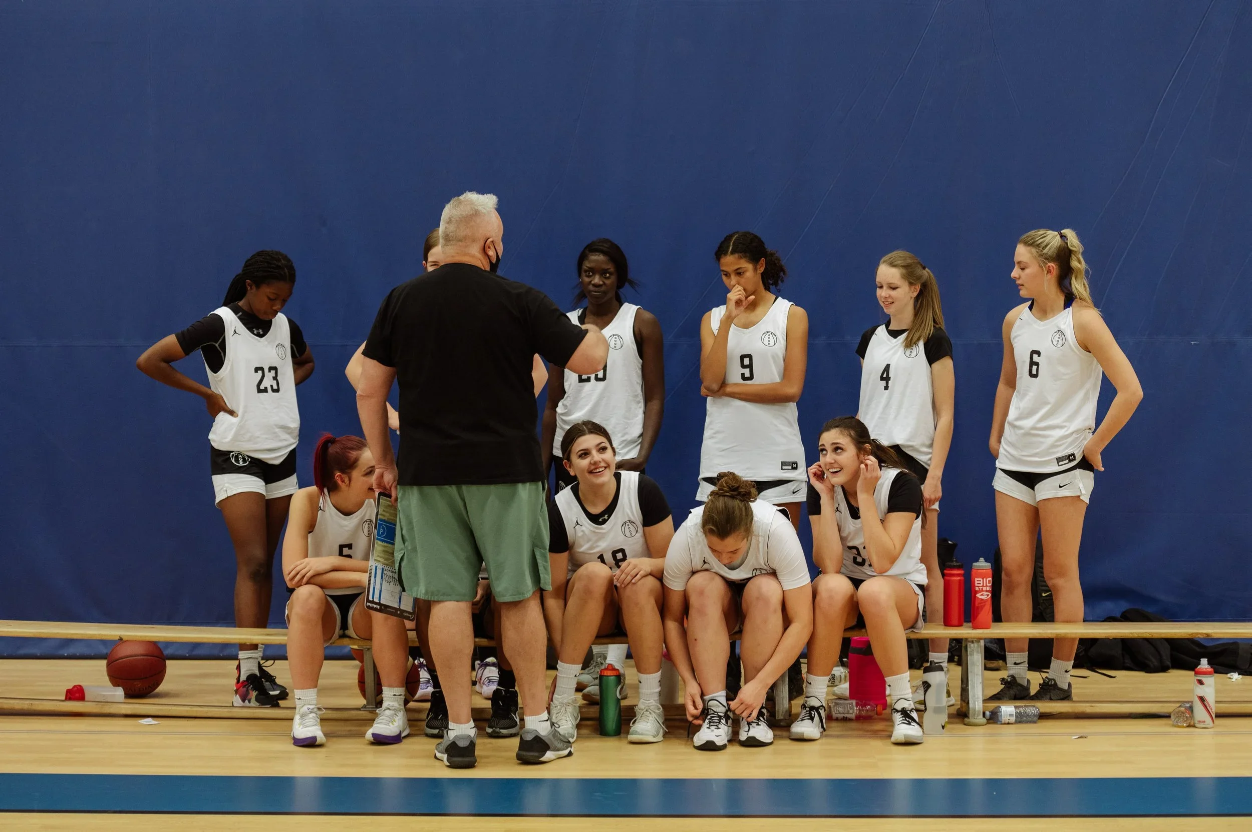 A women's basketball team listening to their coach during a timeout, seated and standing on a gymnasium court.