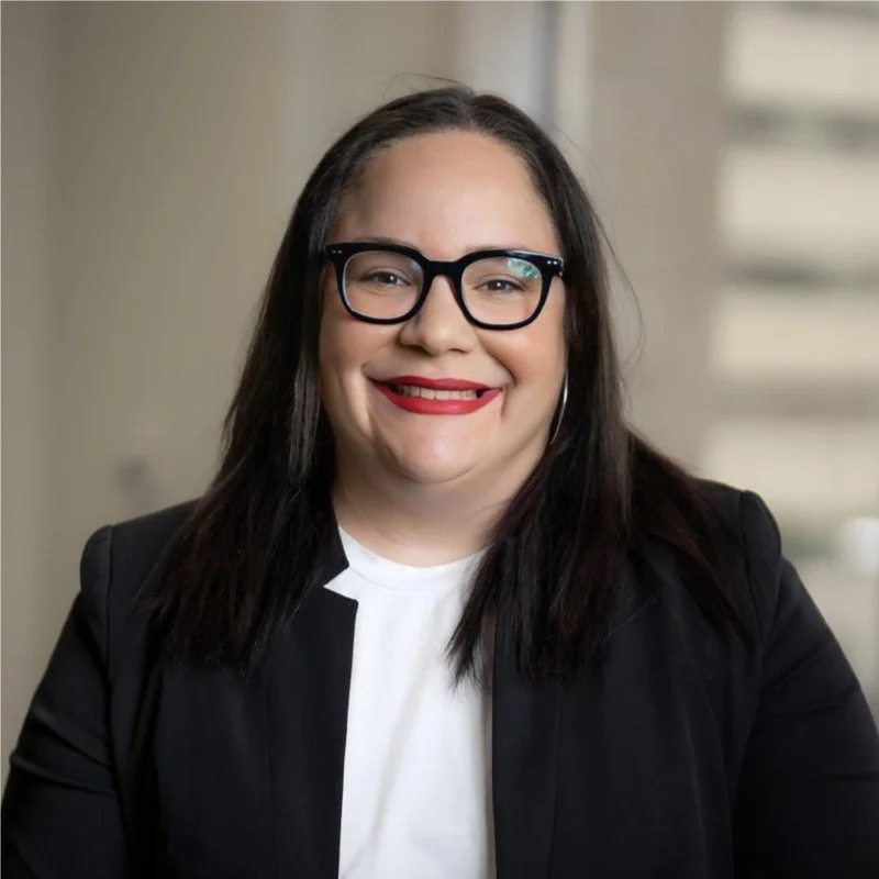 A woman with dark hair, glasses, and red lipstick, smiling in a professional setting.