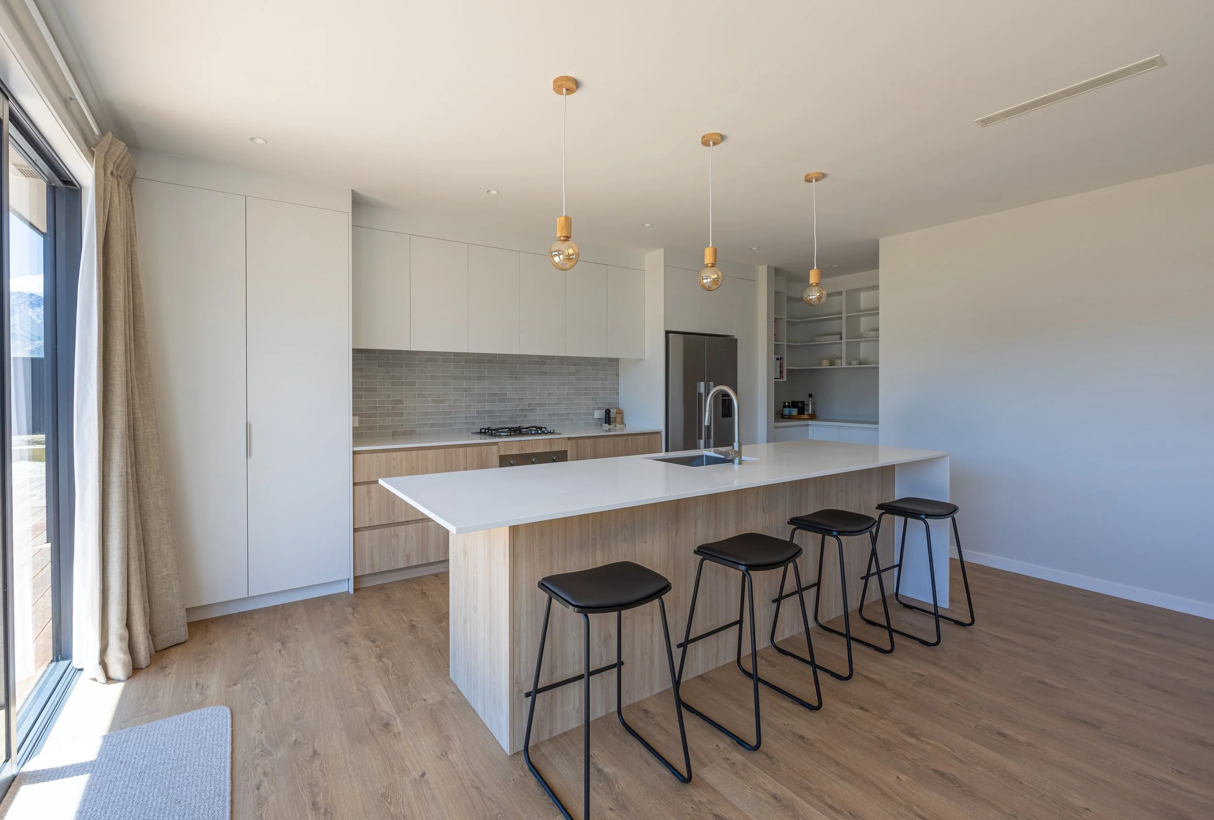 New build in Lake Hāwea, Wānaka. Photo taken looking into the kitchen with a grey splashback, hanging gold lights, white cabinetry