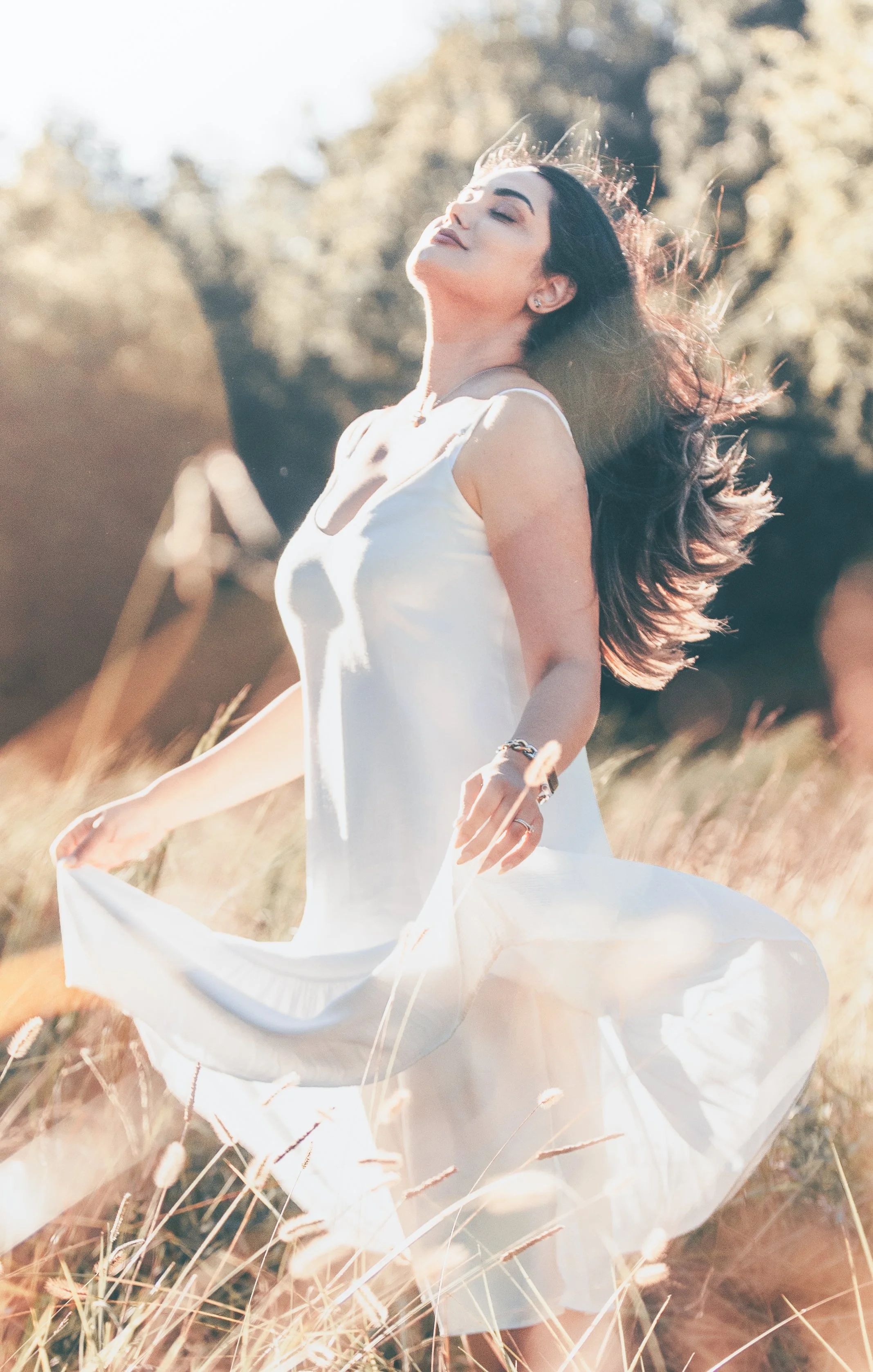 A woman with long dark hair wearing a white dress is standing outdoors in a field of tall grass, with sunlight shining on her face and hair, appearing joyful and relaxed.